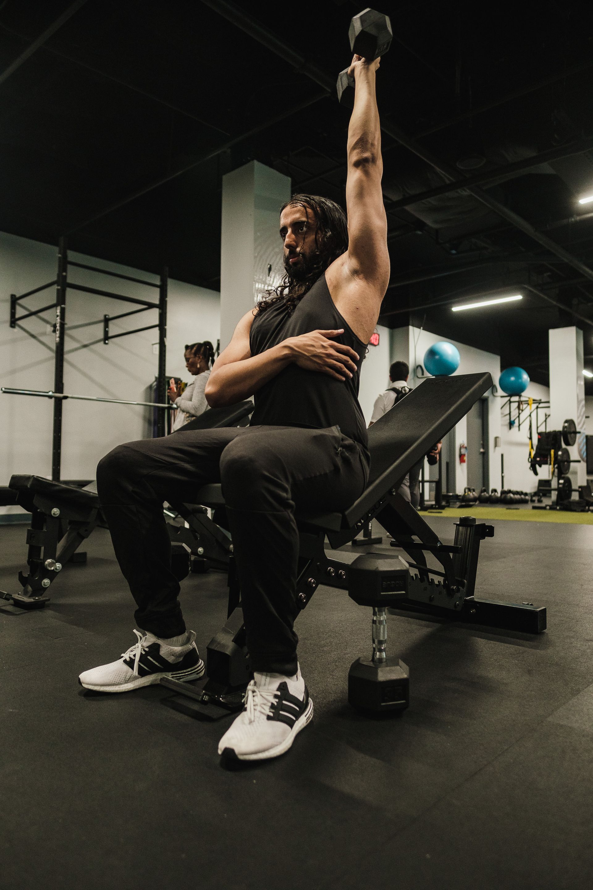 A person performs a single-arm dumbbell overhead press while seated on a weight bench in a gym.