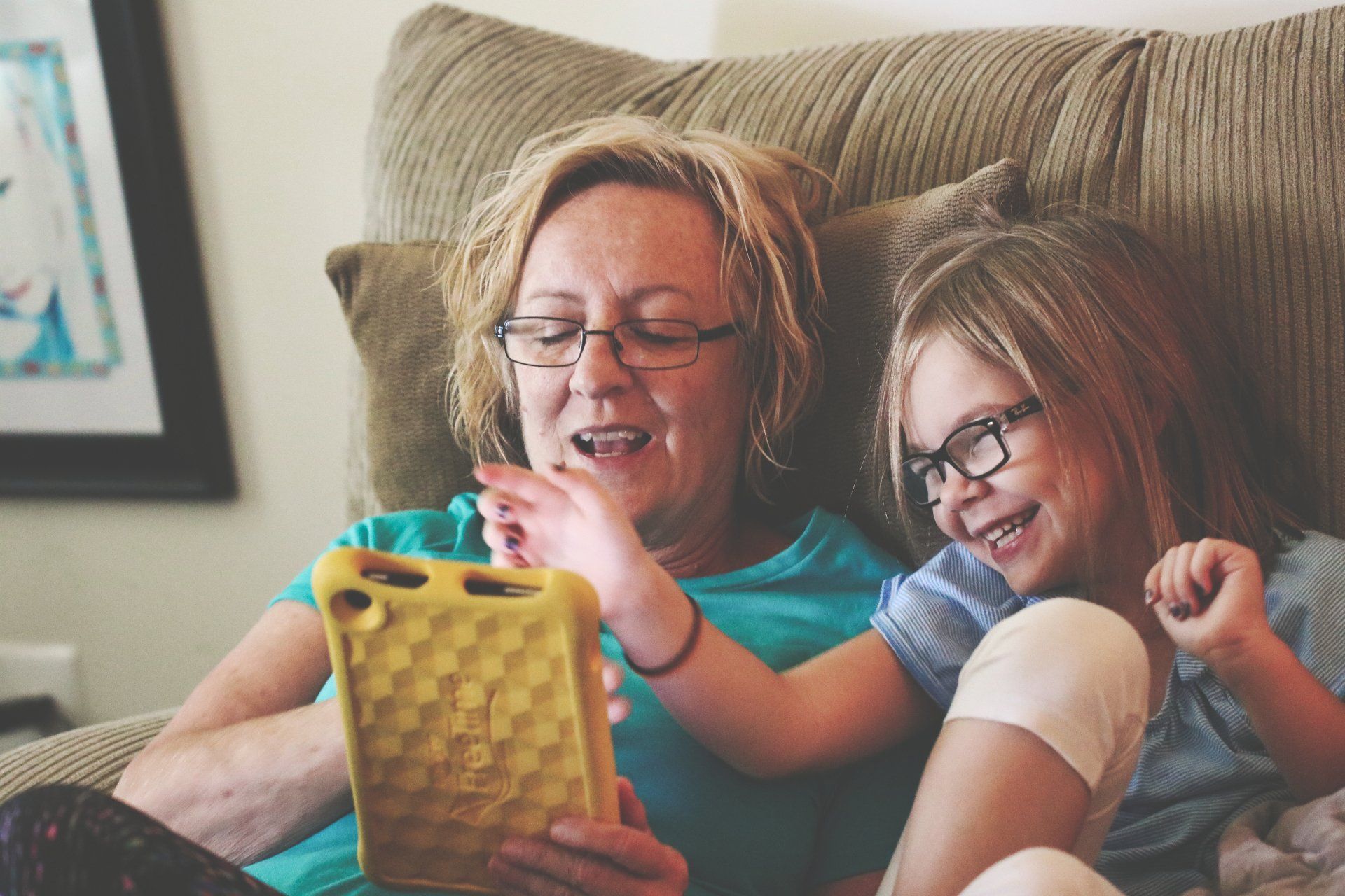 A woman and her young daughter, both wearing glasses smiling and playing on a tablet.