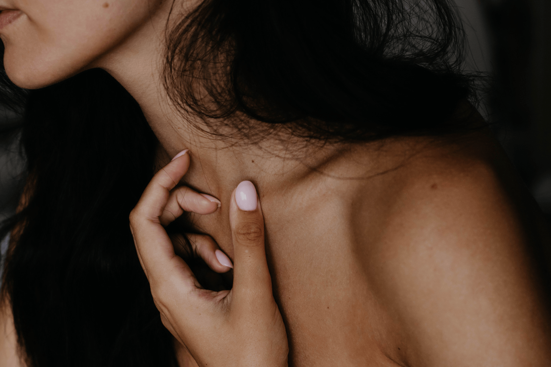 Woman's hand gently touching her neck; dark hair, neutral skin tone, soft lighting.