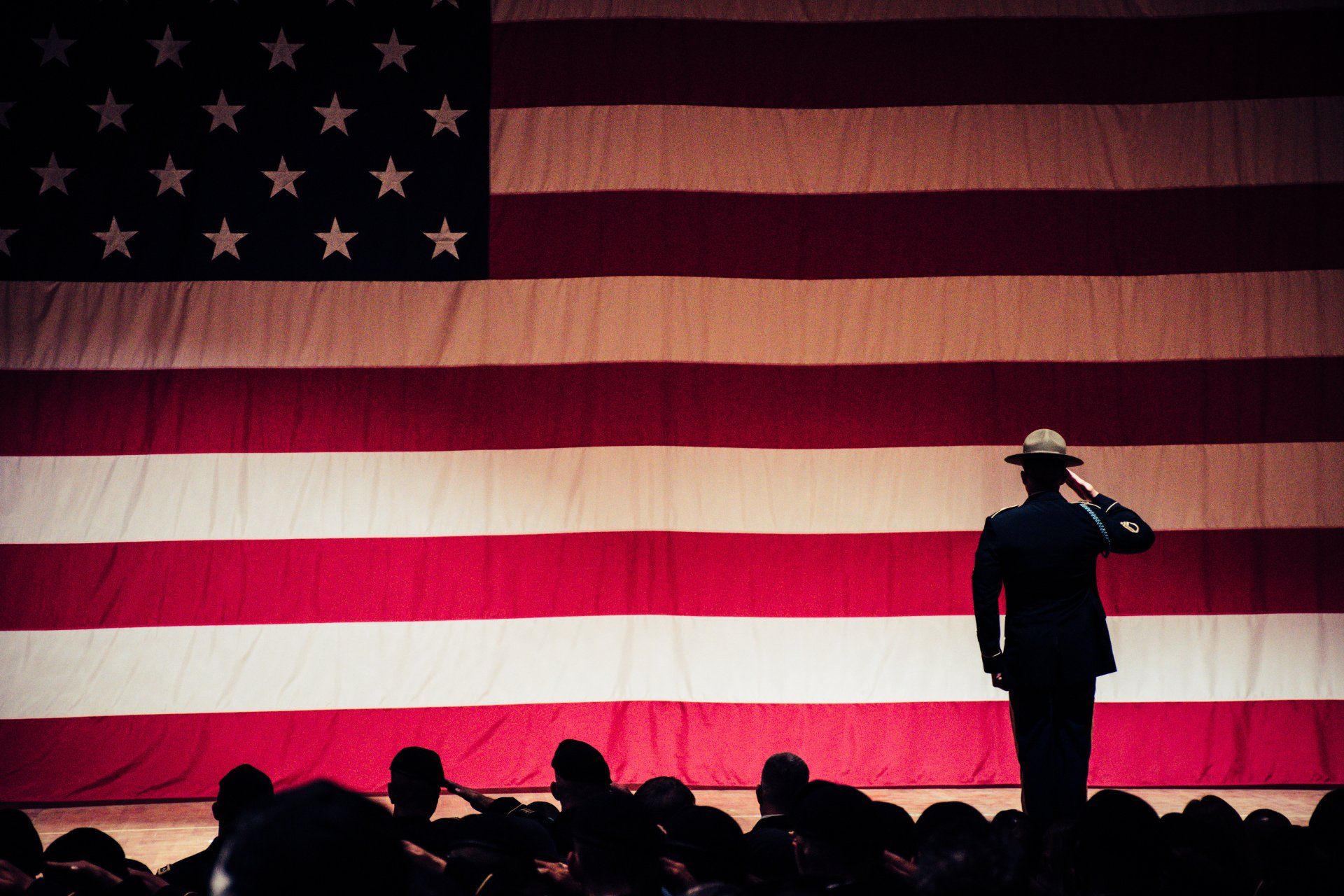 Soldier salutes in front of a giant American flag, with other silhouettes saluting in the audience.