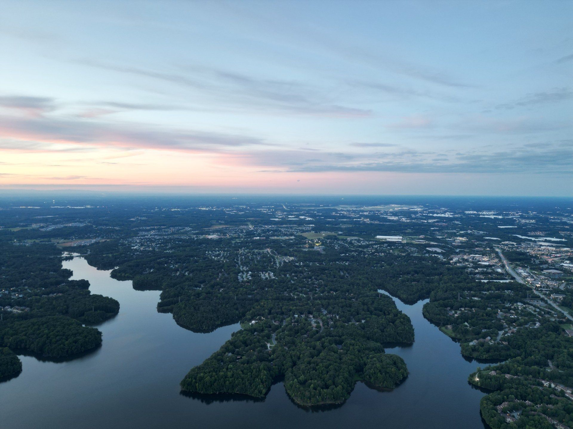 Aerial view of a lake with islands and surrounding cityscape under a pastel sunset sky.