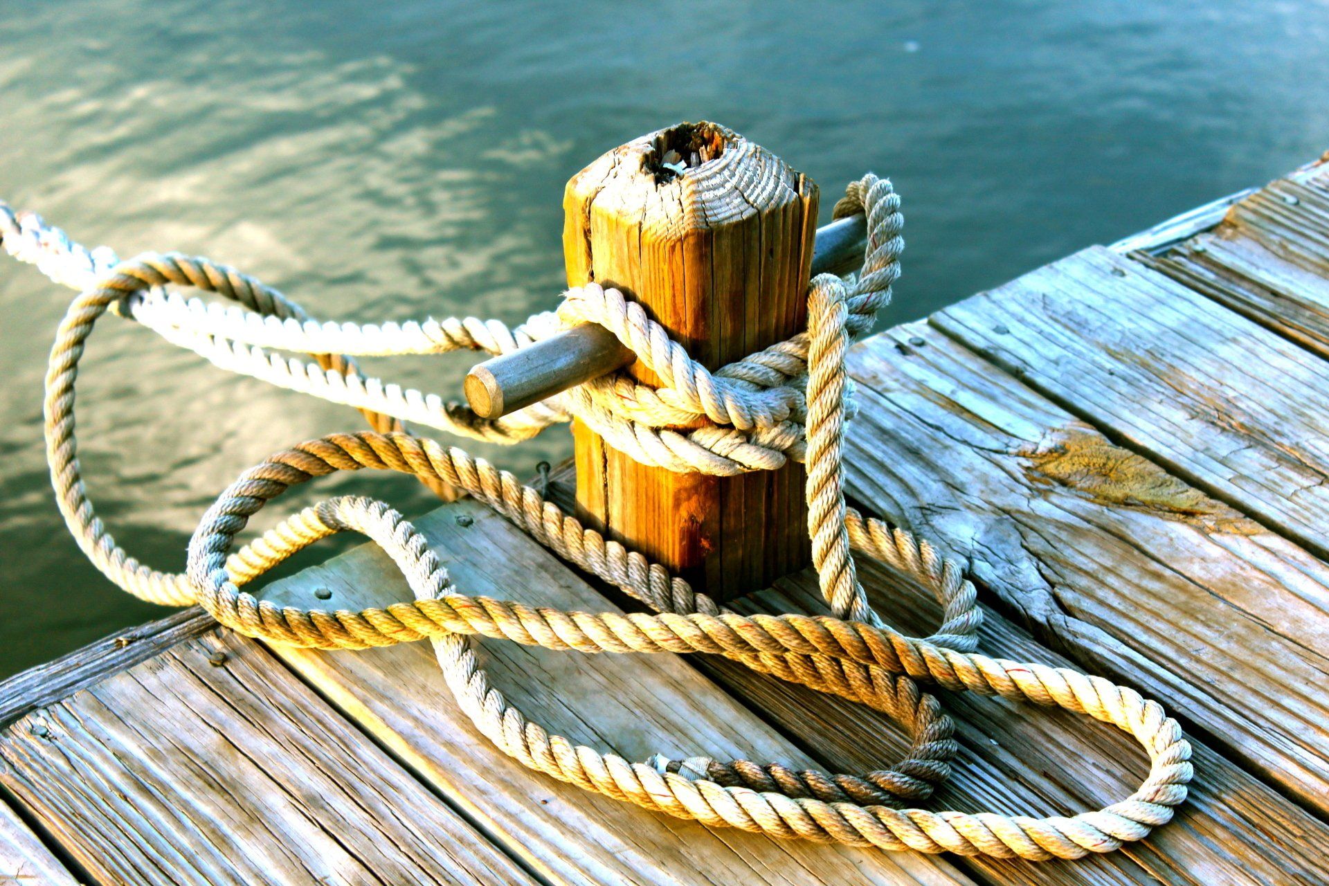 A thick, weathered rope is coiled and secured around a wooden mooring post on a wooden pier overlooking the water.