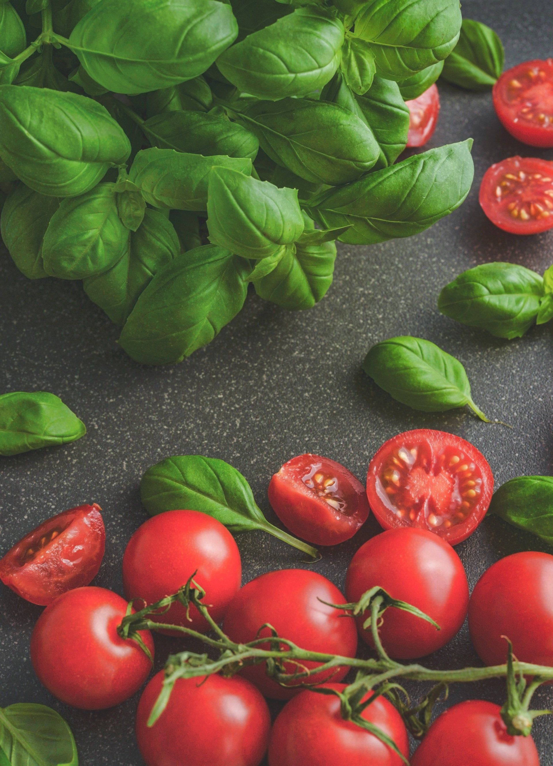 Fresh basil leaves and red cherry tomatoes, some whole and some halved, arranged on a dark, textured surface.