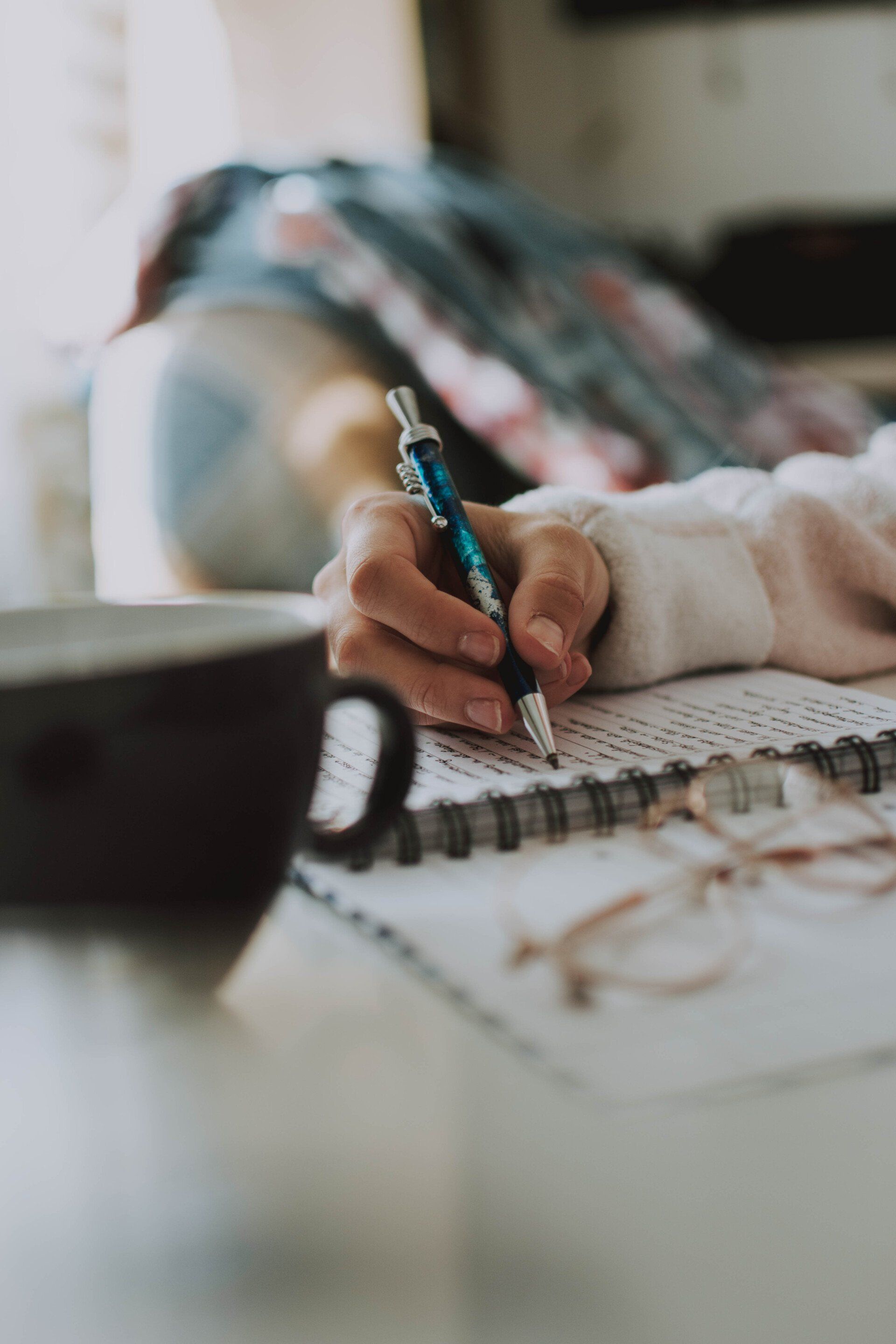 Picture A person is writing in a notebook next to a cup of coffee.