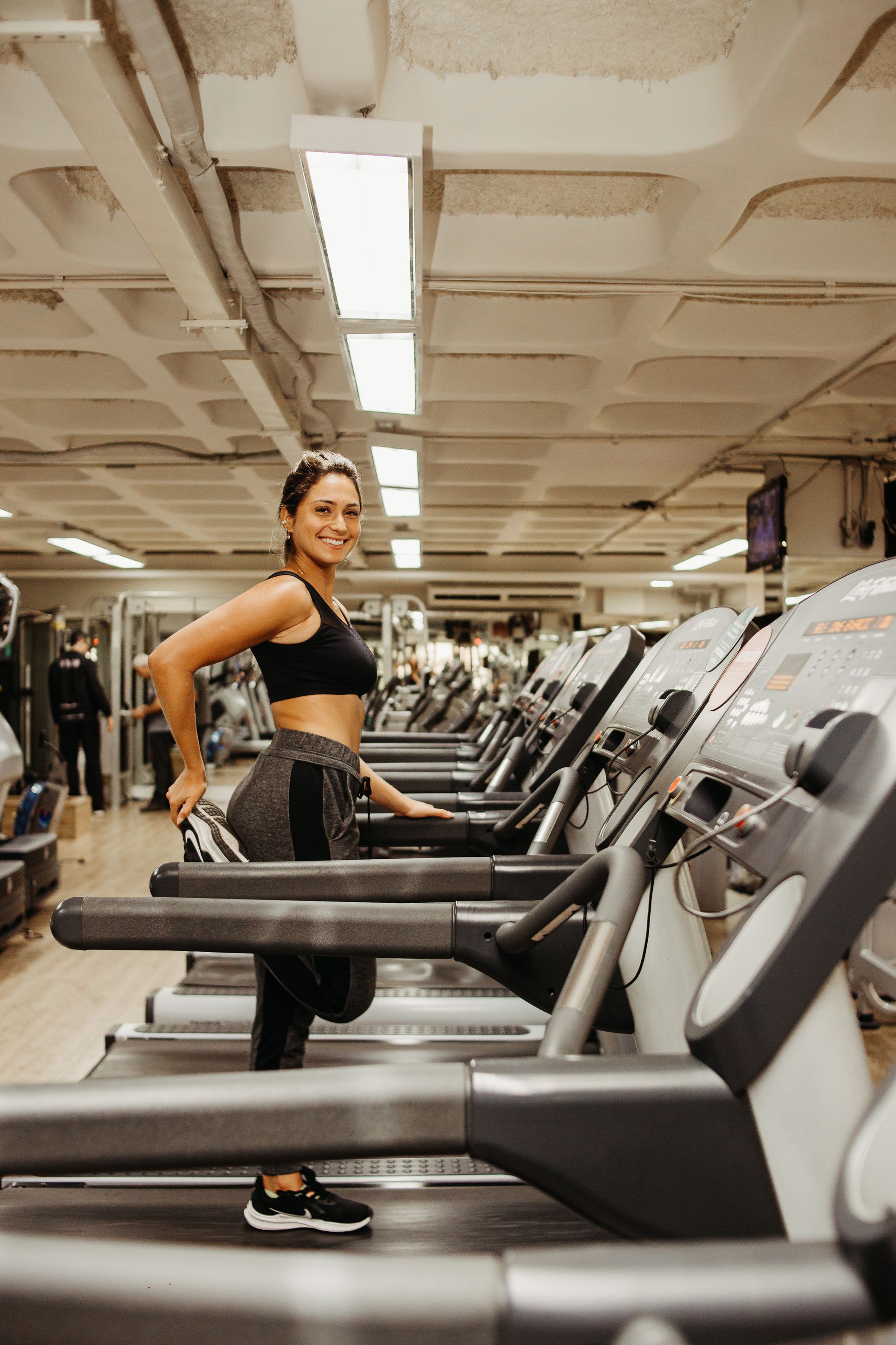 Mulher em pé ao lado de esteiras em uma academia, vestindo roupas de ginástica pretas e sorrindo.