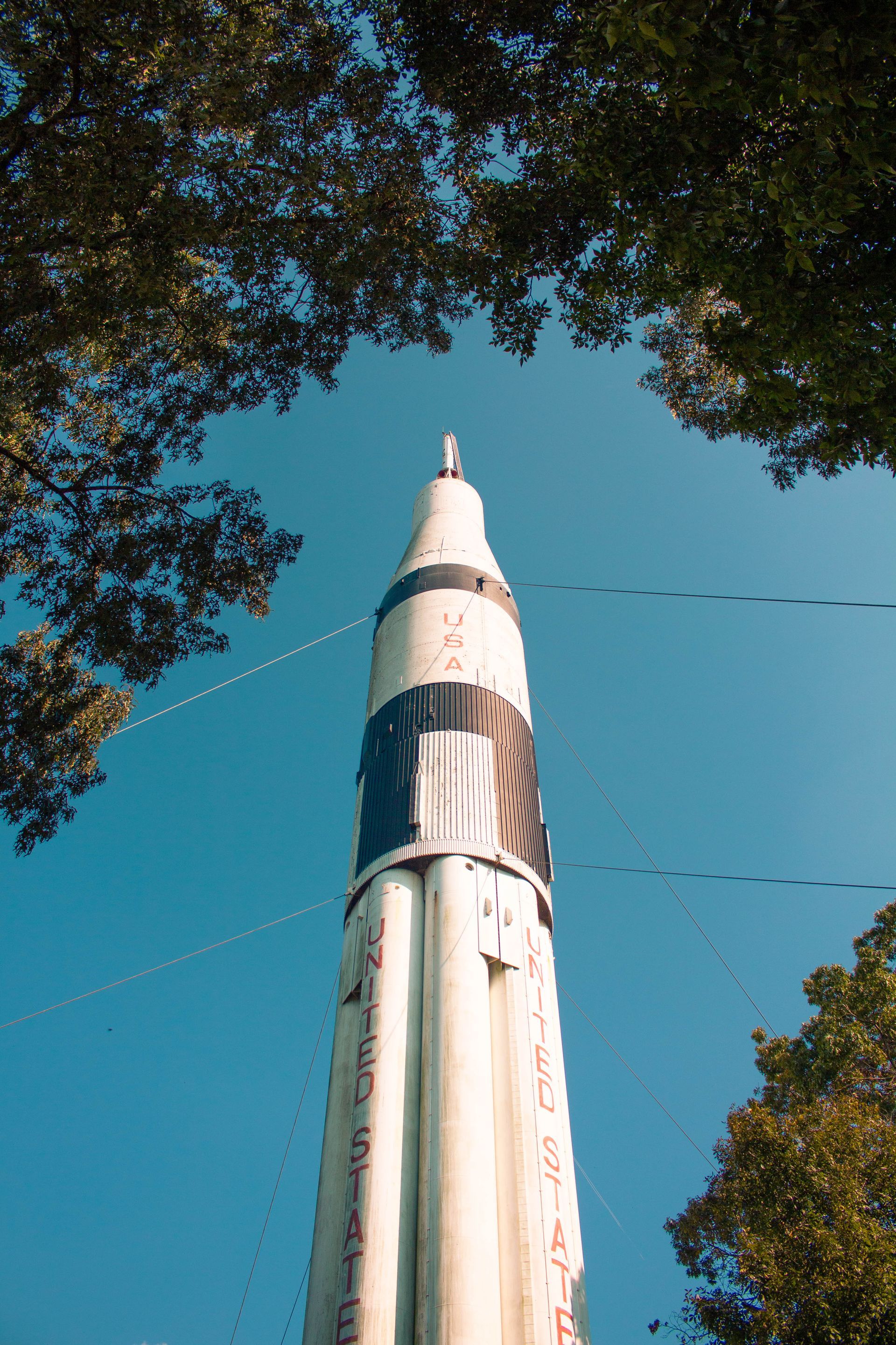 Tall white rocket seen from below against a blue sky, framed by trees