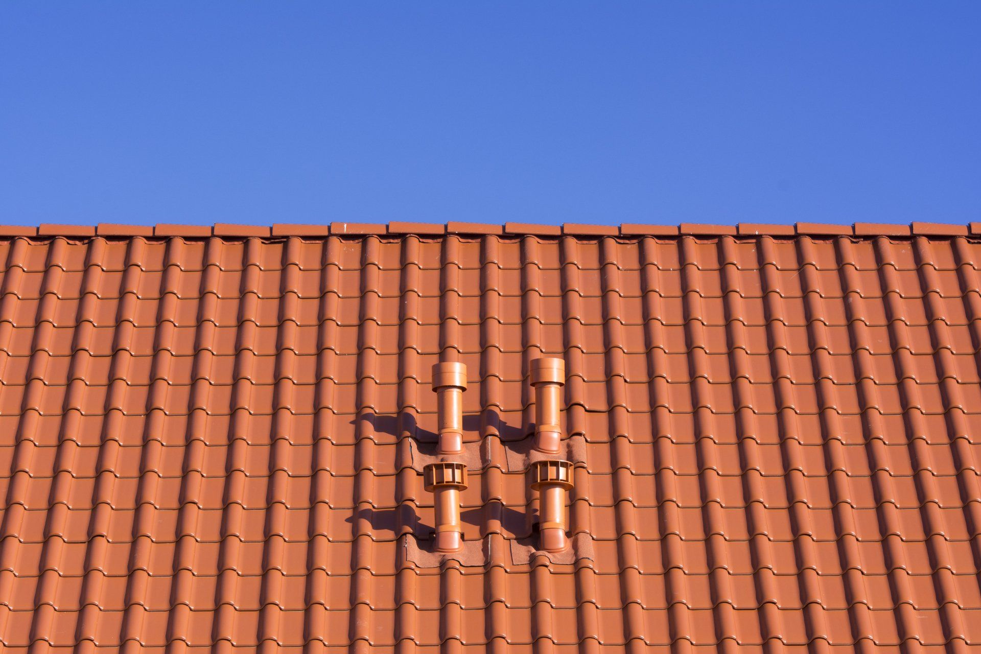 Red tiled roof with two brown pipes against a clear blue sky.