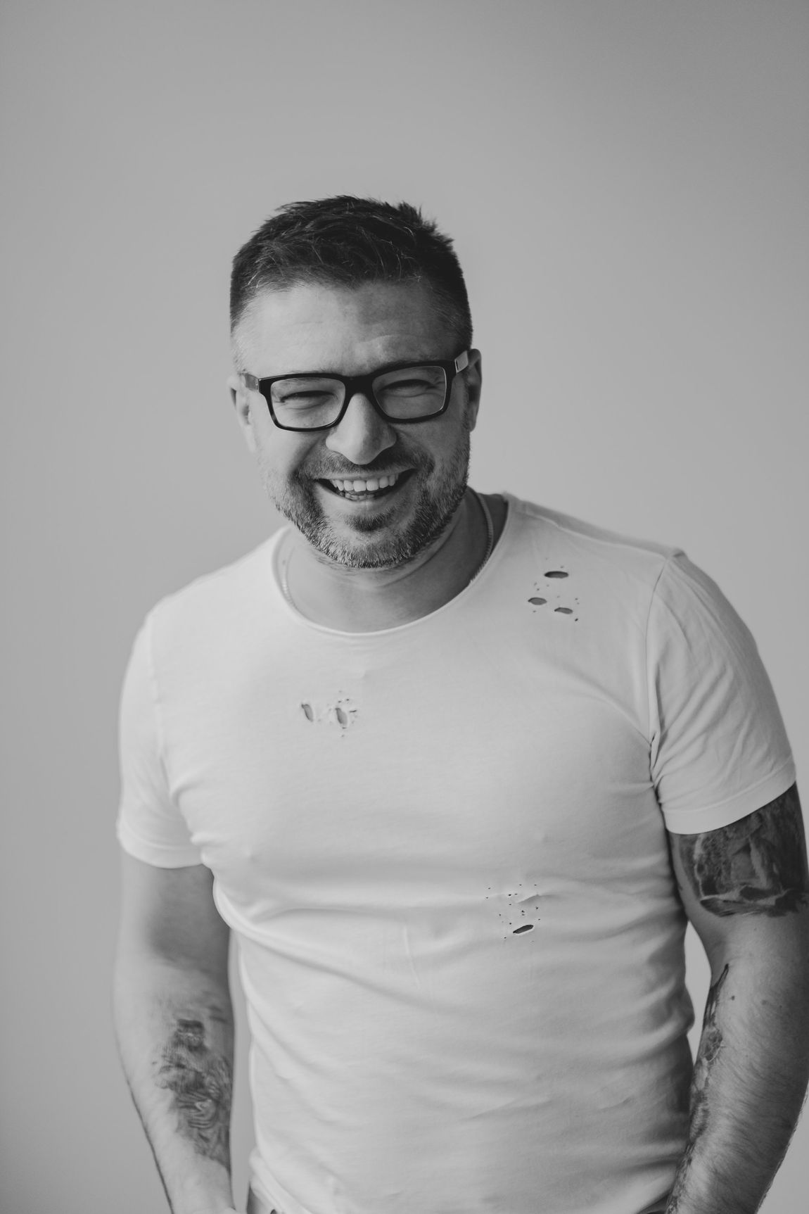 Man with glasses smiles, wears a white t-shirt. Gray background. Tattoos visible on arms.