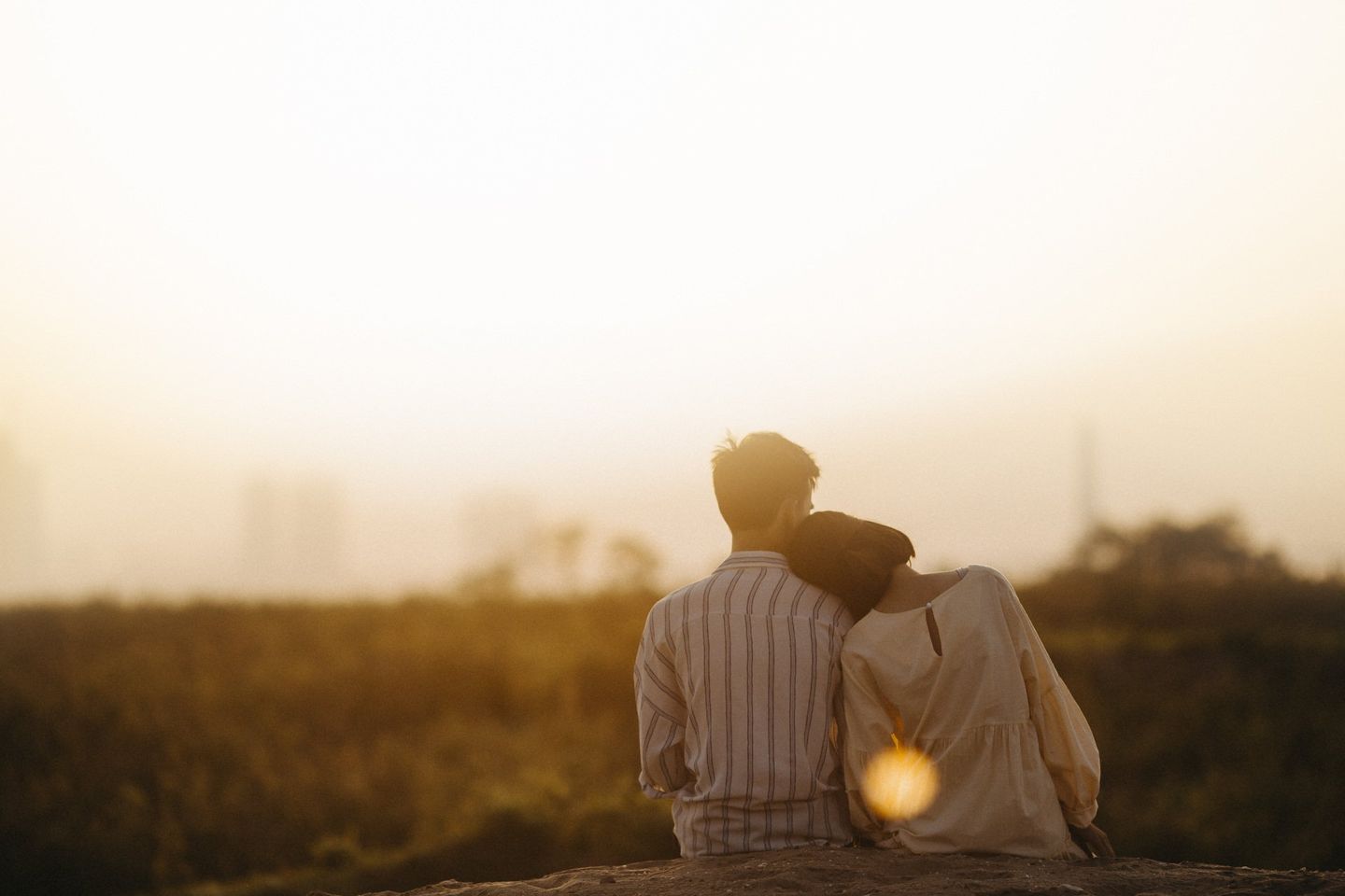 A man and a woman are sitting on top of a hill looking at the sunset.