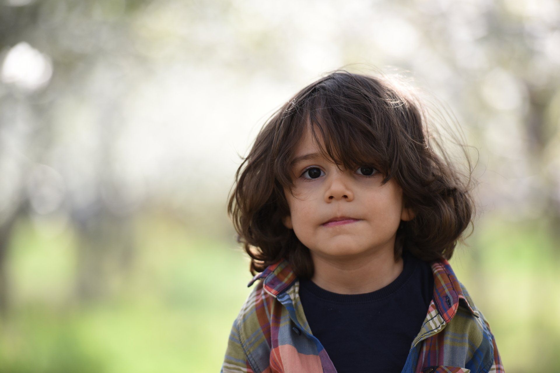 A young boy with long hair is wearing a plaid shirt and looking at the camera.