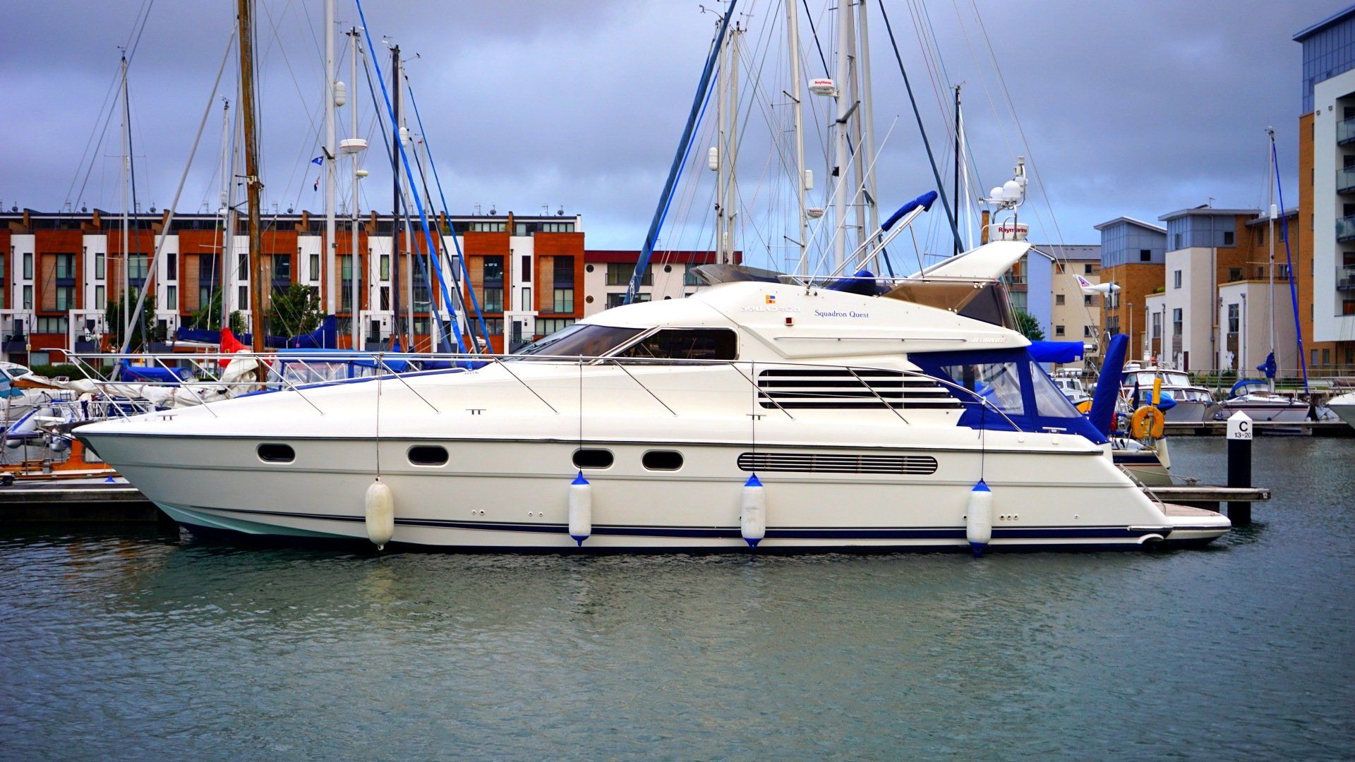 White motor yacht docked in marina, with buildings and sailboats in background.