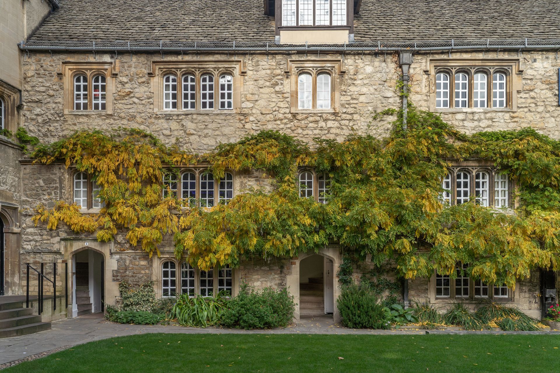 A historic stone building with mullioned windows, draped in lush green and yellow vines, facing a grassy courtyard.