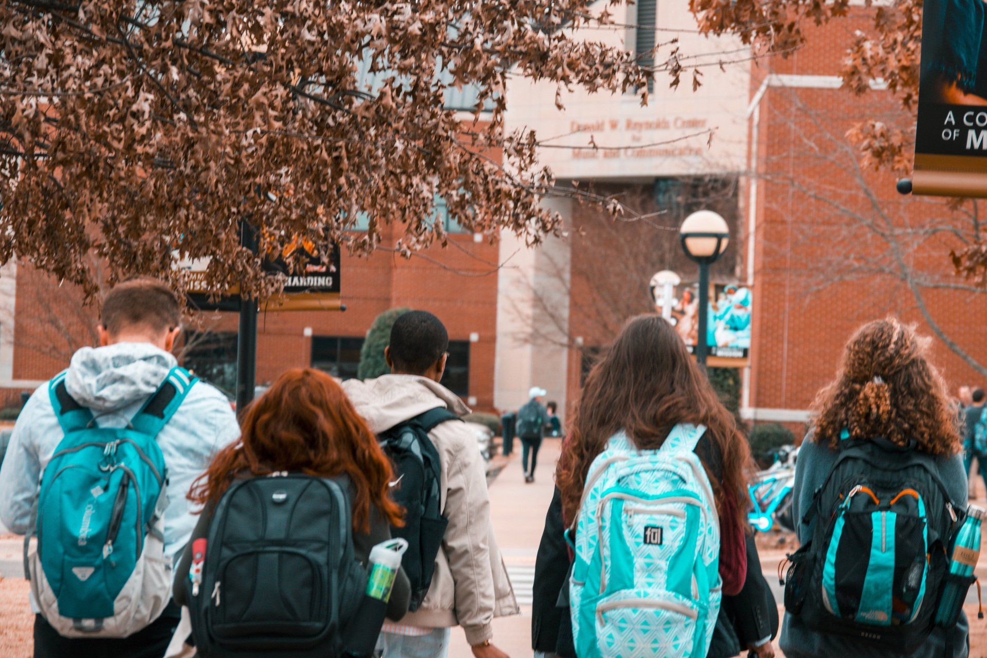 Four students with backpacks walking along a tree-lined campus path toward a brick building