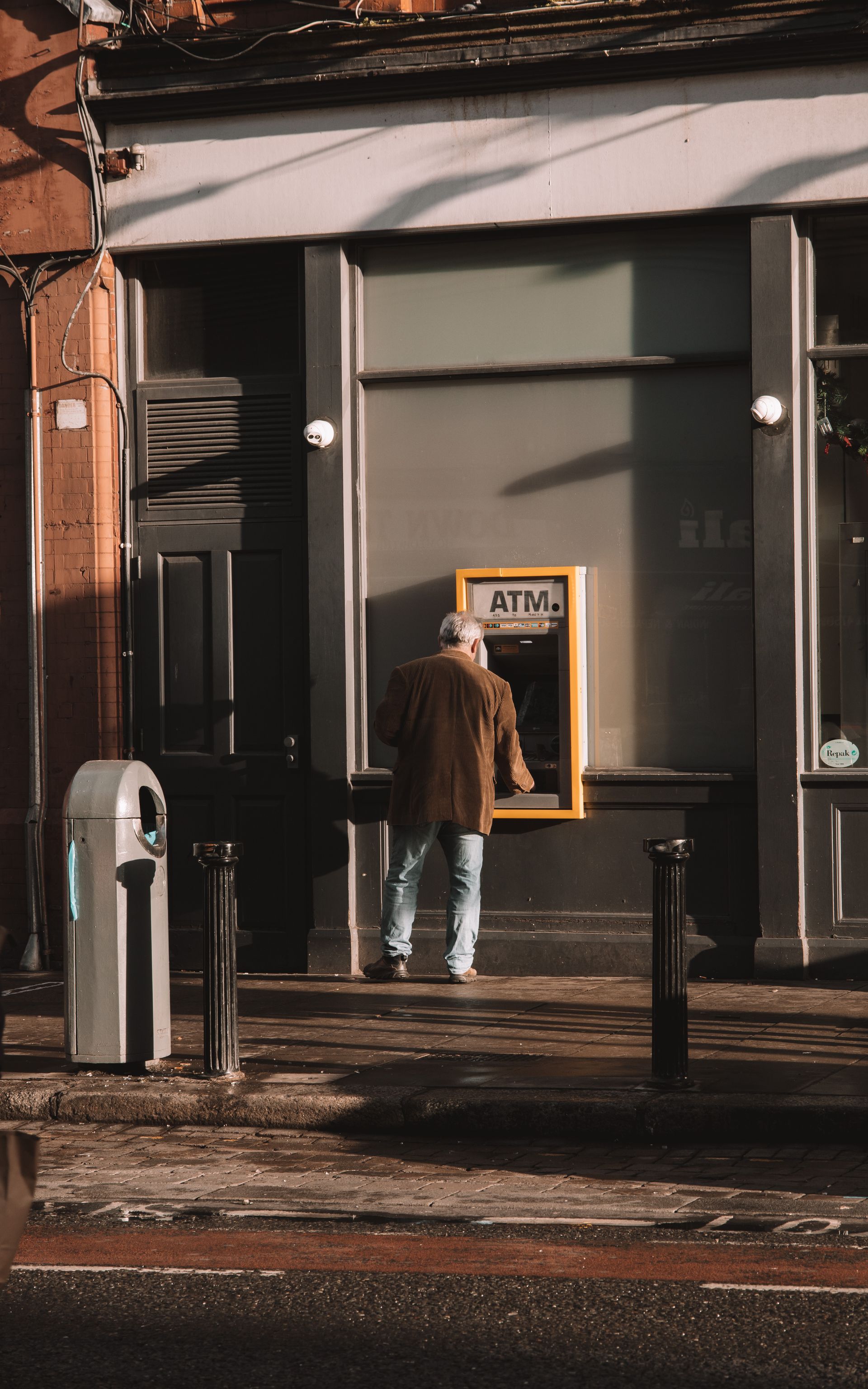 Person using ATM on a city street. Brown jacket, blue jeans. ATM is gold-colored.