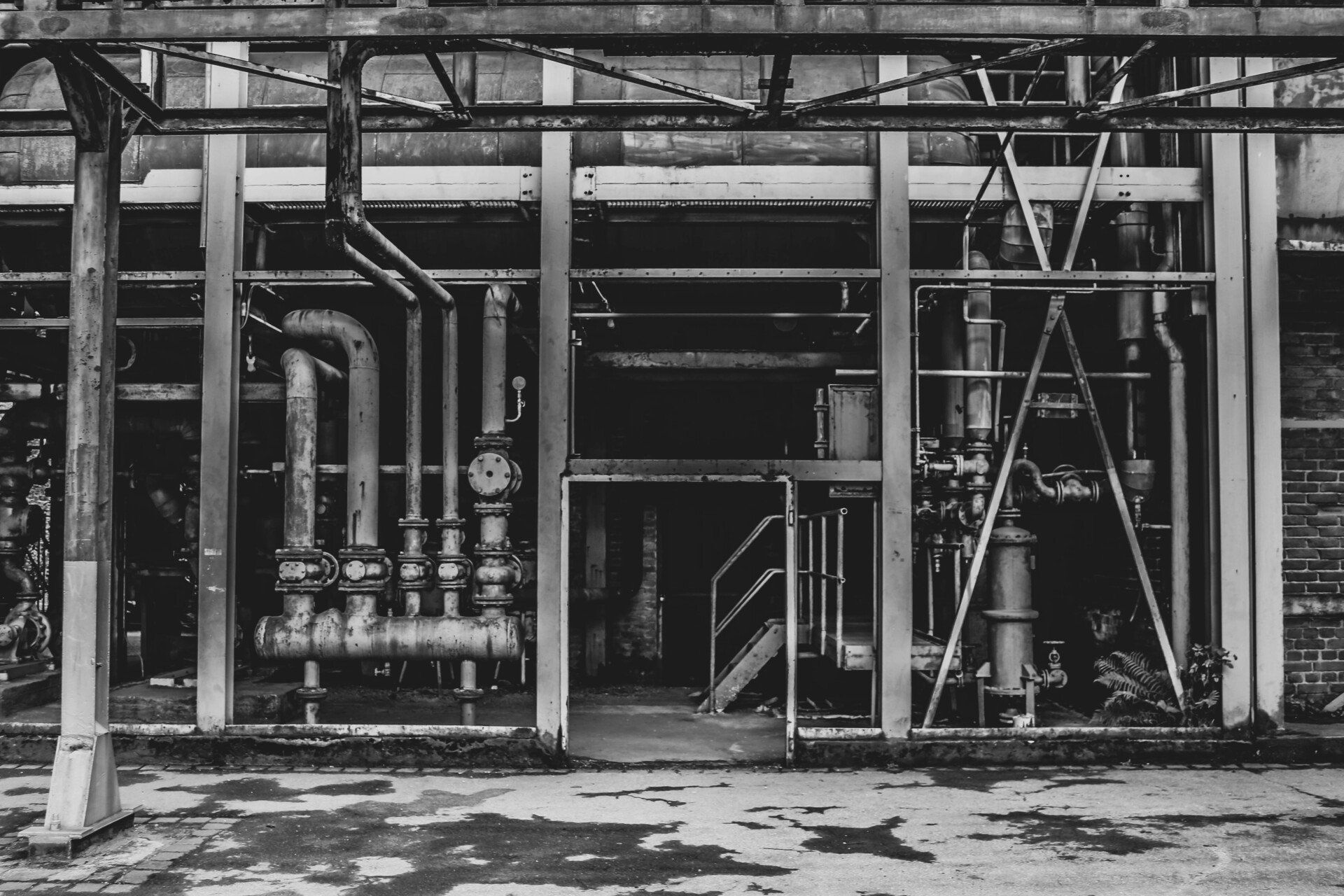 Black and white photo of an industrial setting. Pipes, metal scaffolding, and a doorway.