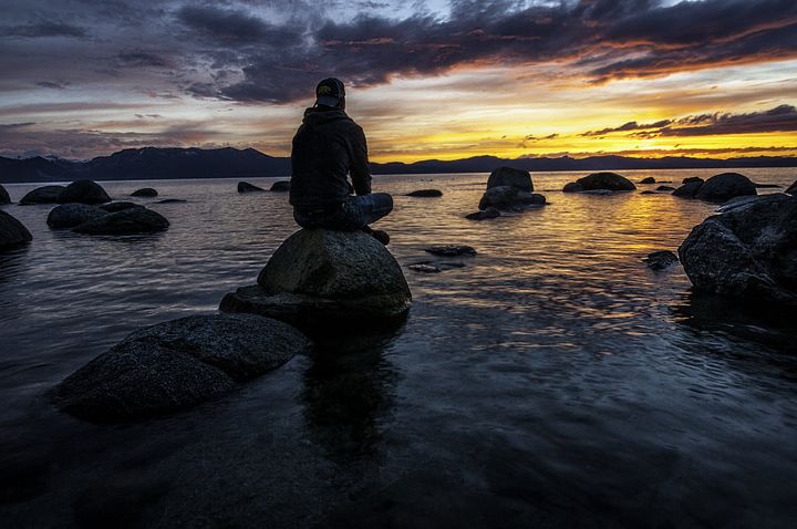 A man is sitting on a rock in the middle of the ocean at sunset.