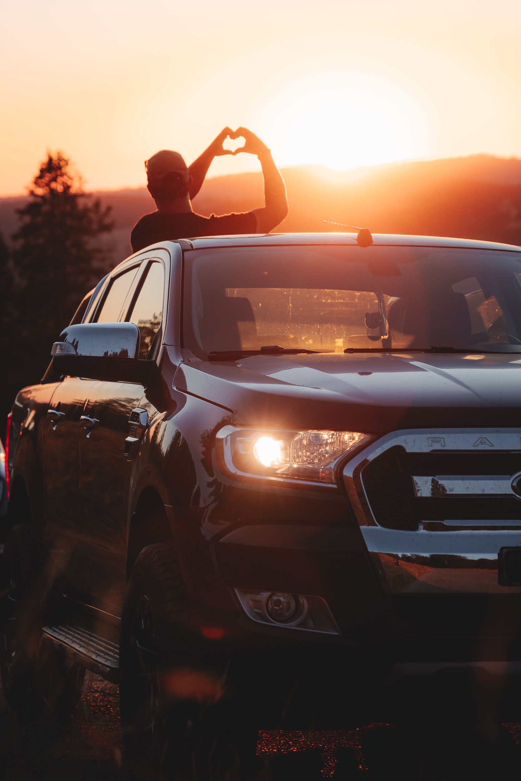 Person on car roof makes heart shape with hands at sunset.