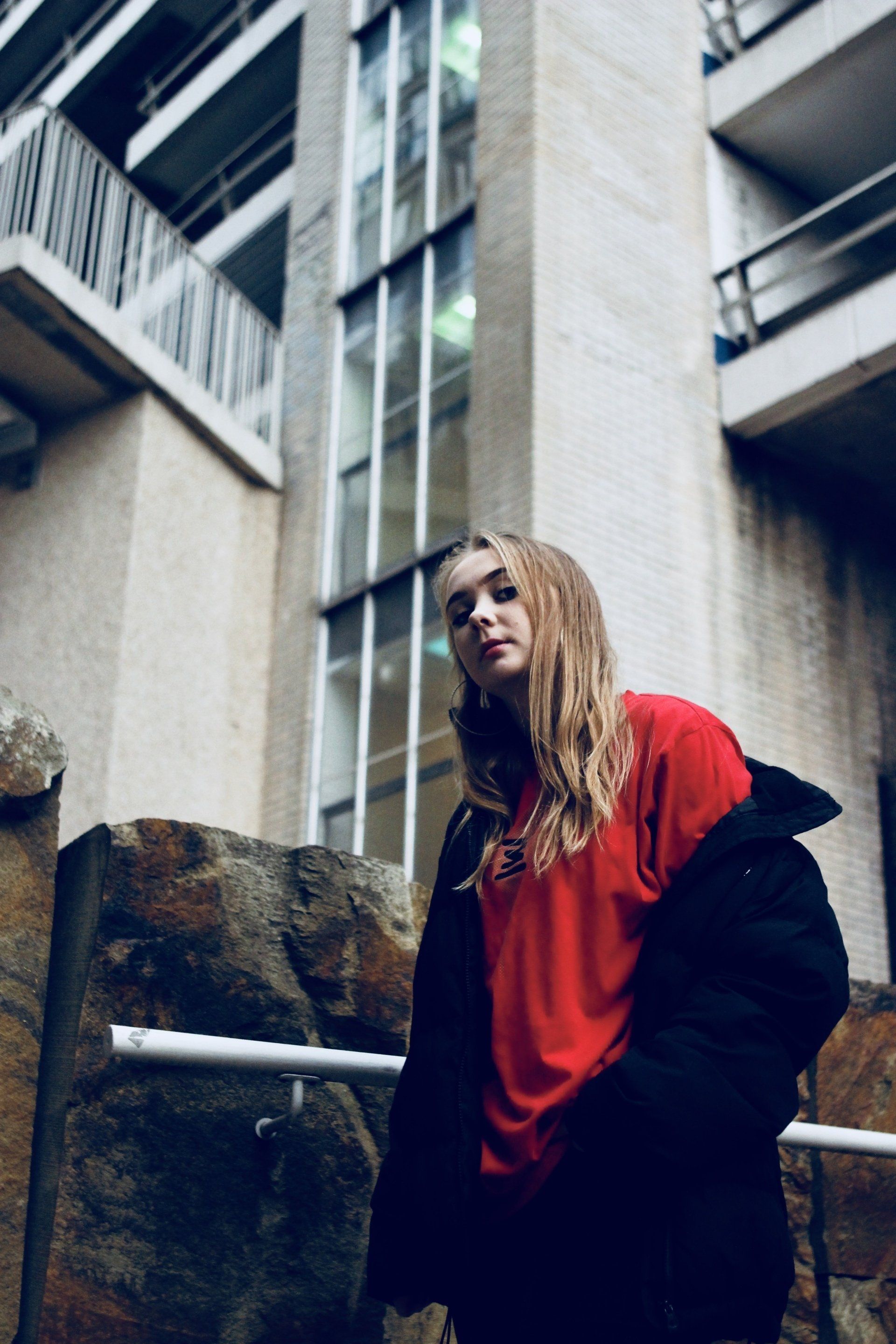 A woman in a red shirt and black jacket is standing in front of a building.