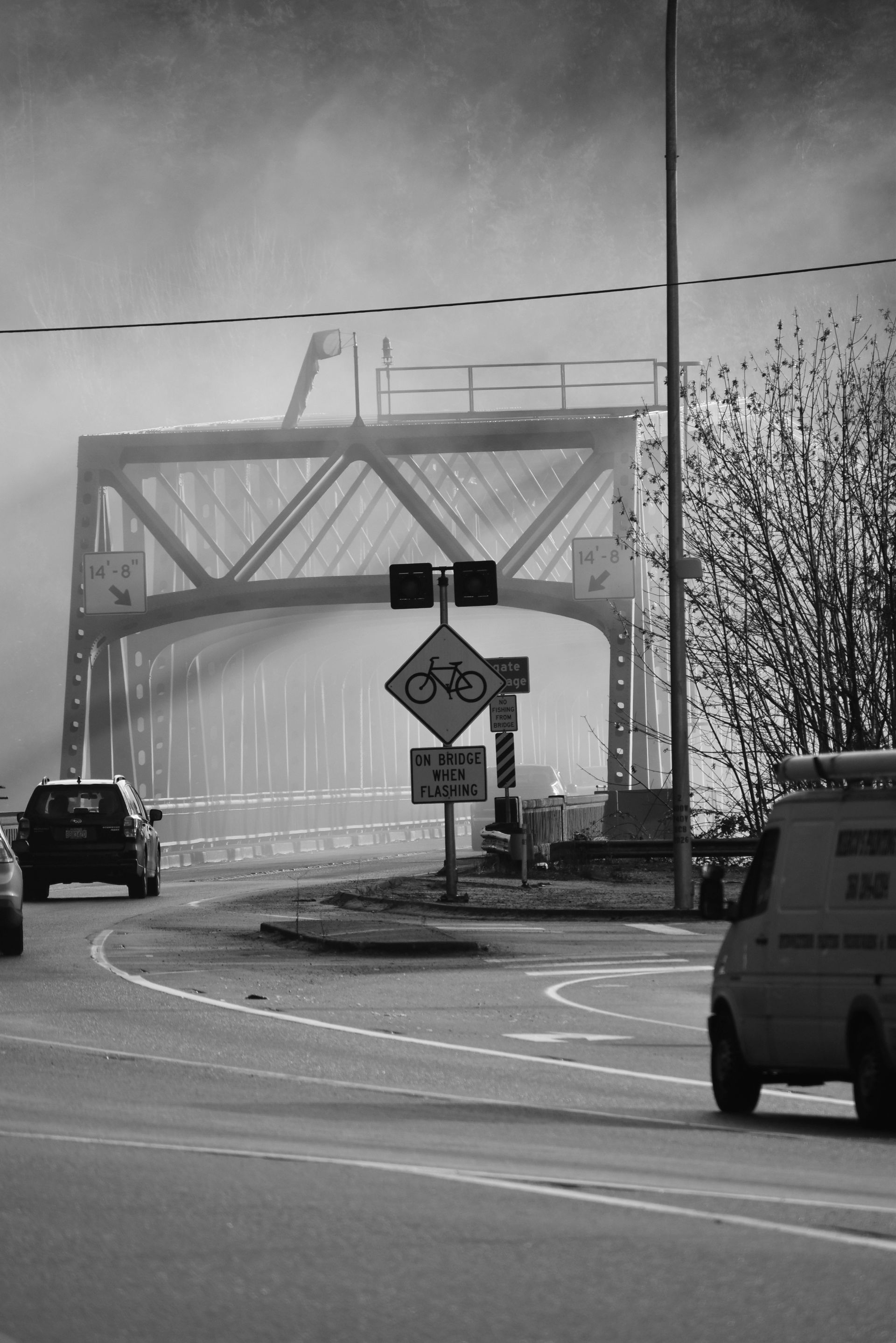 Foggy road leading to a bridge, with cars and traffic signs in grayscale