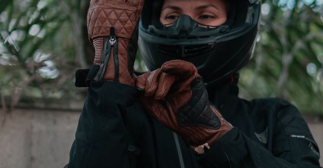 Person in a black motorcycle helmet and jacket adjusting brown leather gloves outdoors.