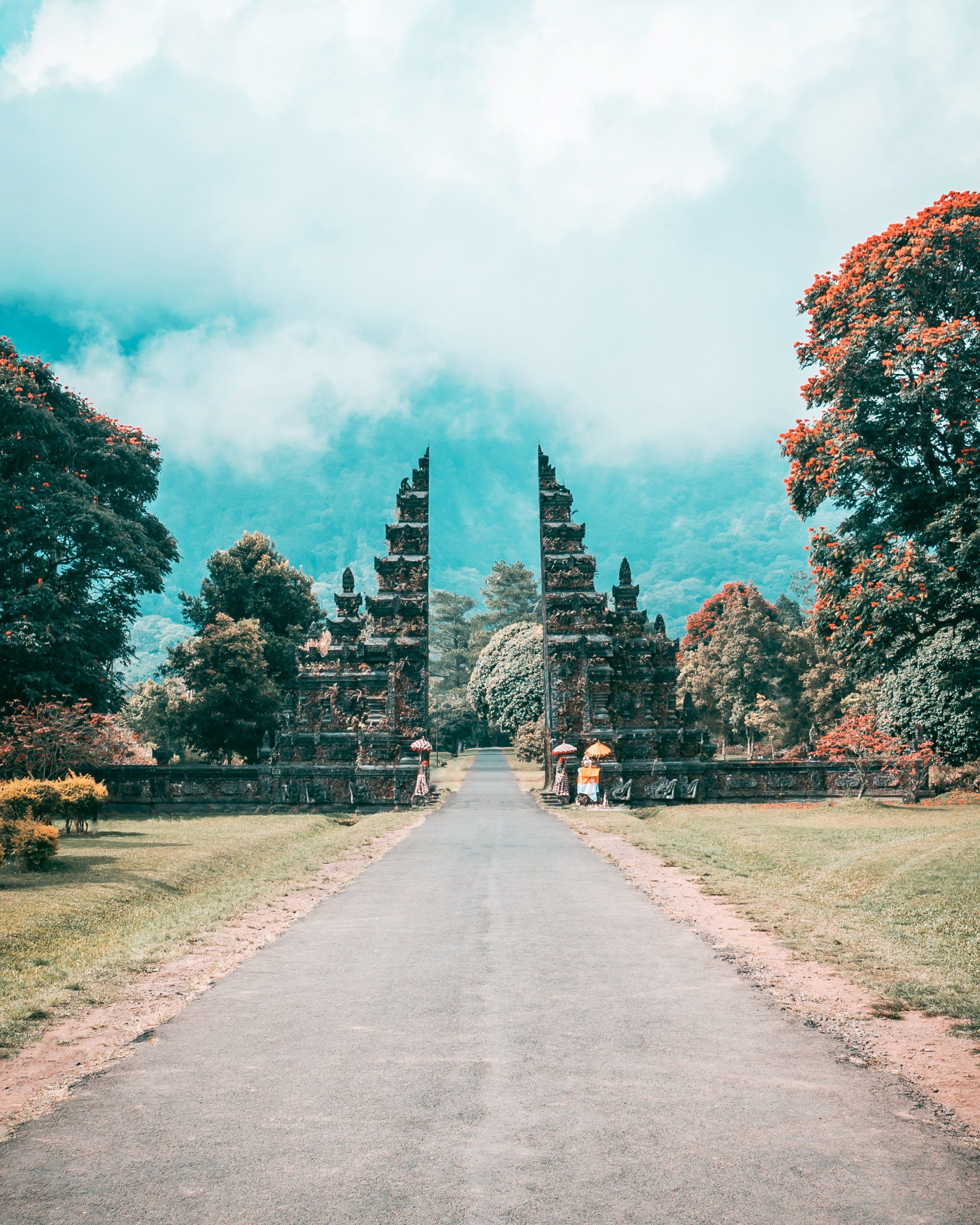 Stone gate leads to a path, surrounded by trees and lush greenery against a cloudy, teal sky. Bali