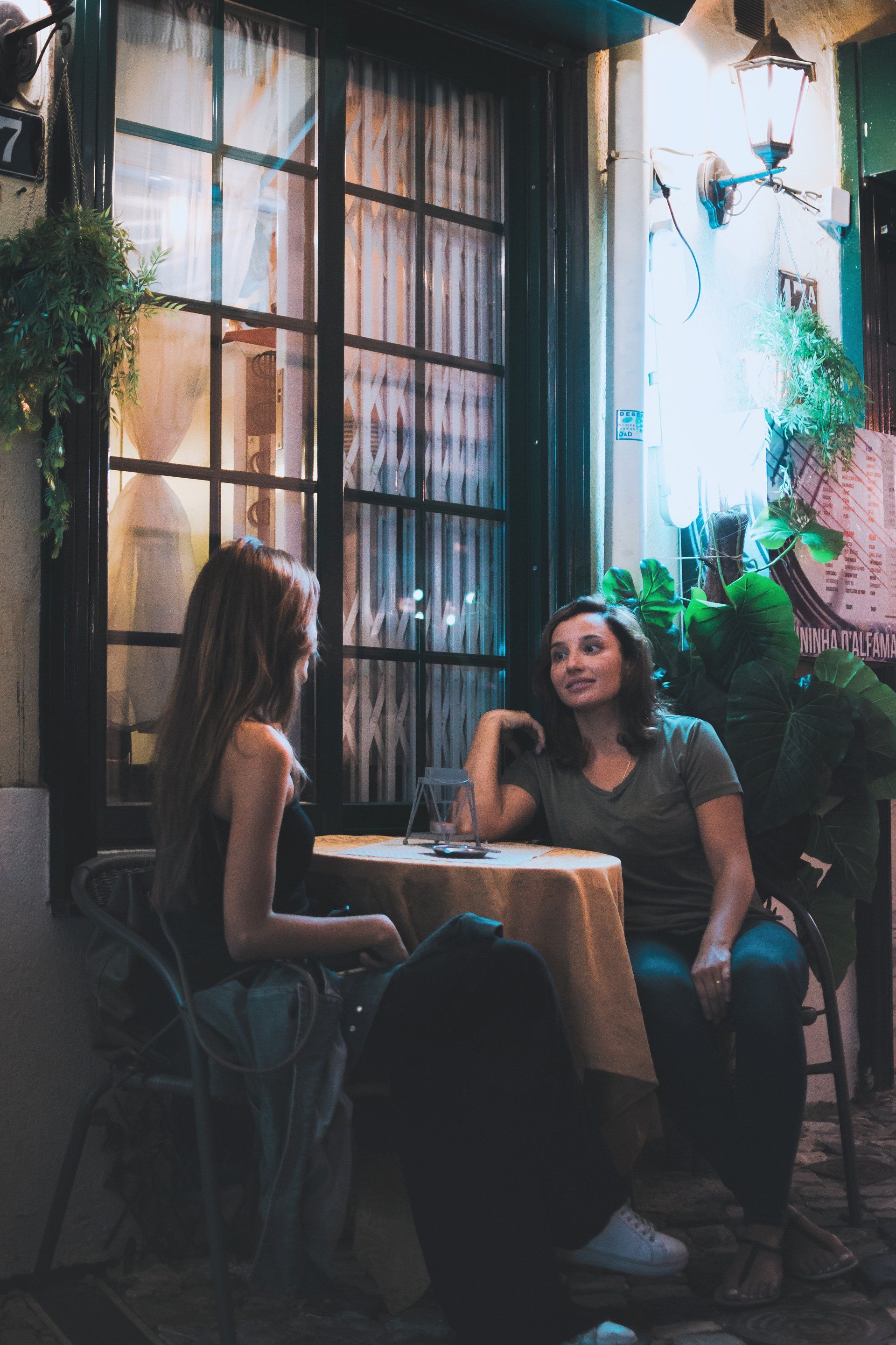 Two women are sitting at a table outside a restaurant talking to each other.