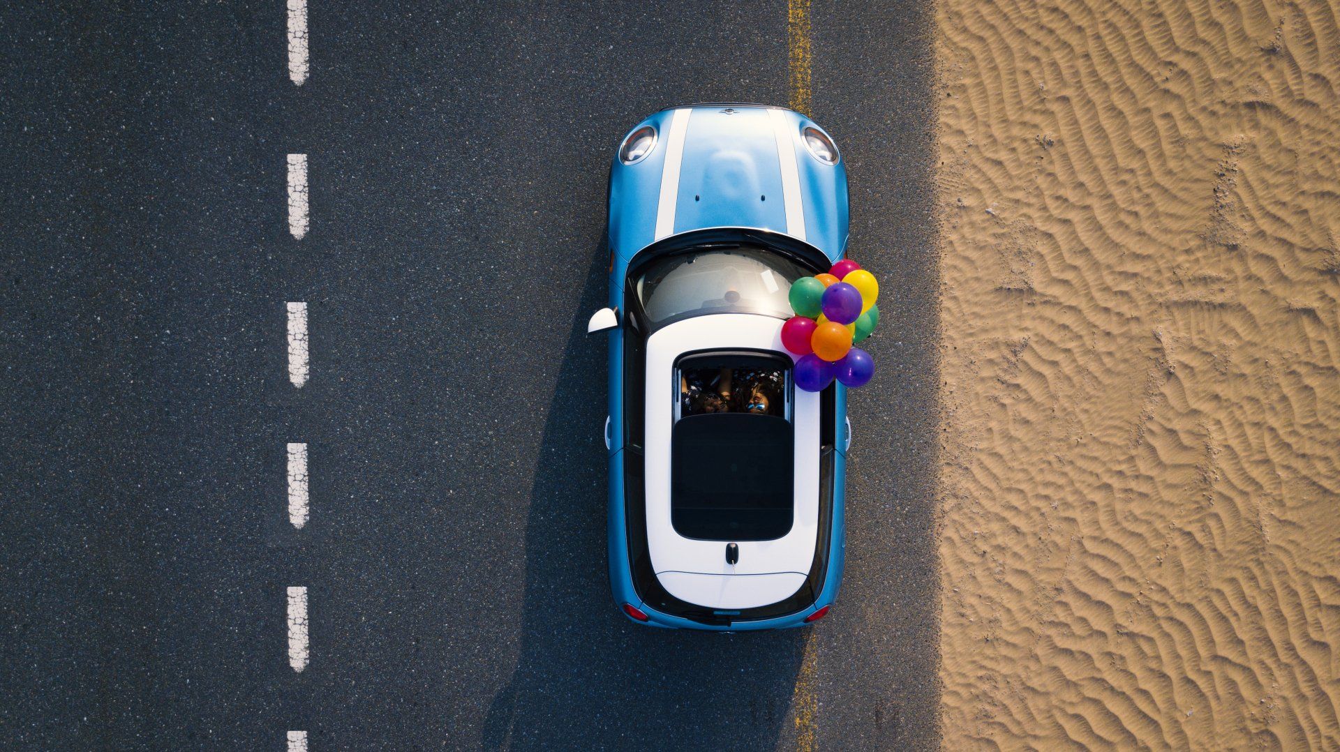 An aerial view of a blue car with balloons in the back driving down a road.