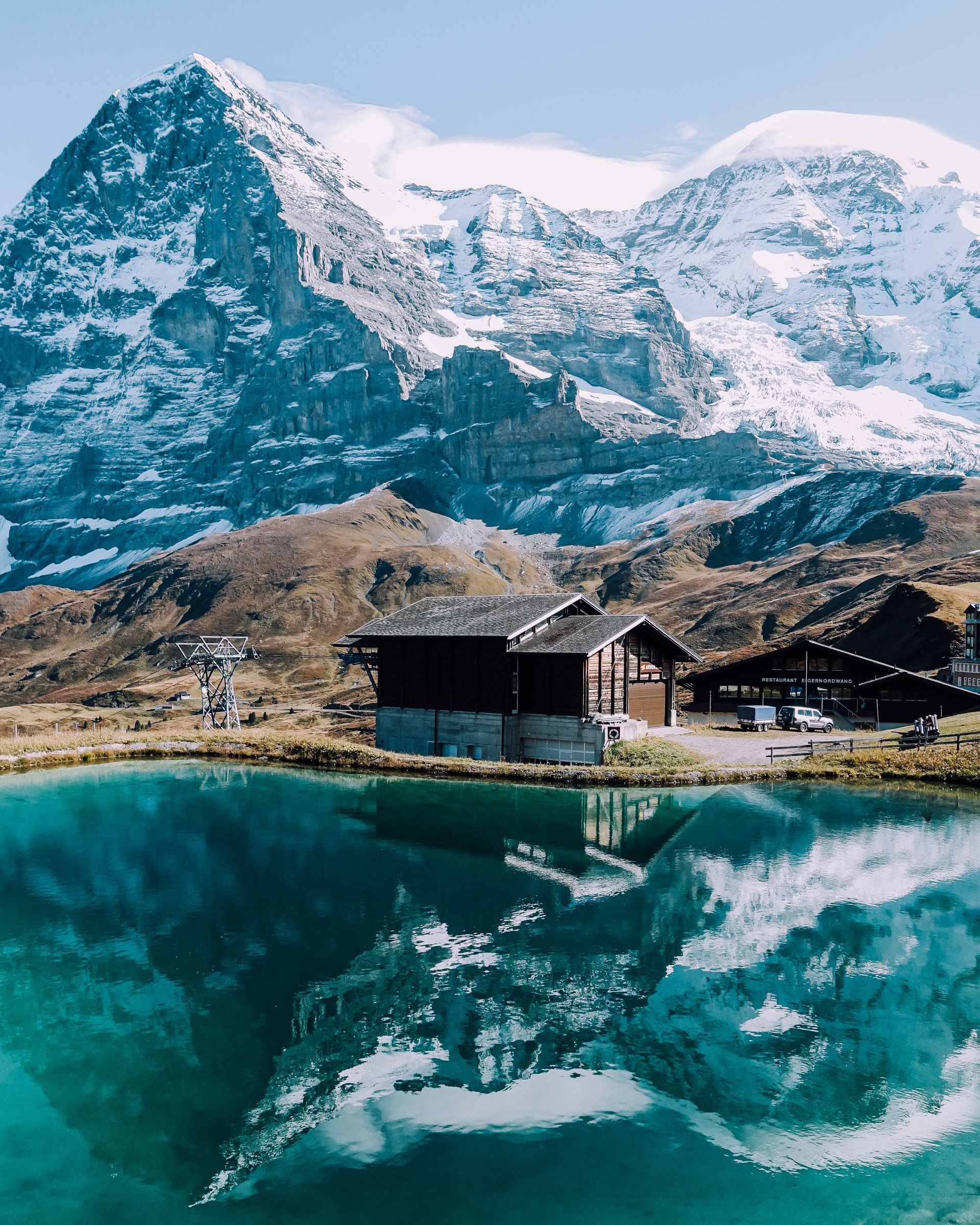 A rustic wooden cabin sits beside a vibrant turquoise lake, reflecting the dramatic, snow-capped Swiss Alps in the distance.