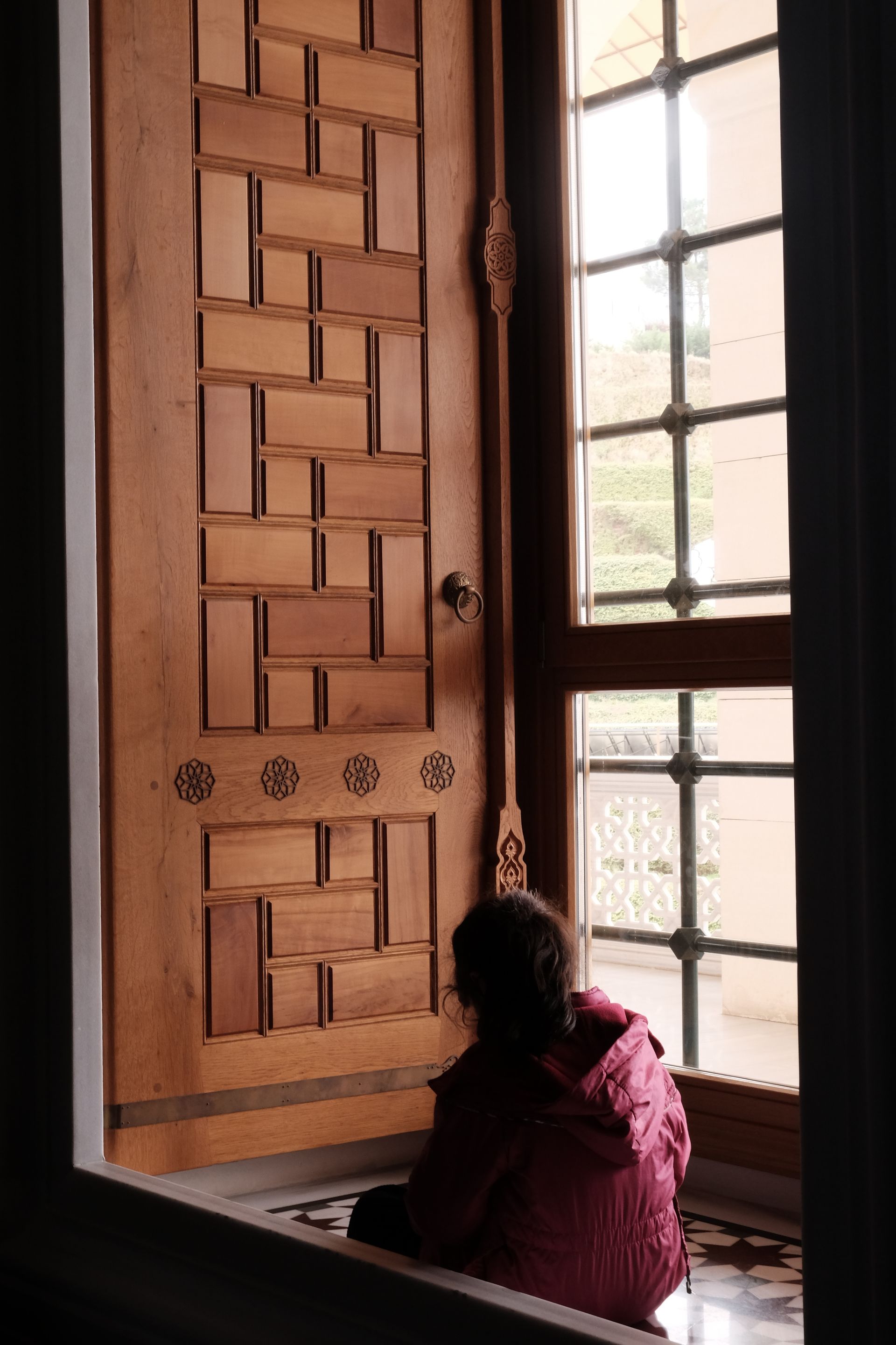 Child in red jacket sits by window, looking at ornate wooden door.