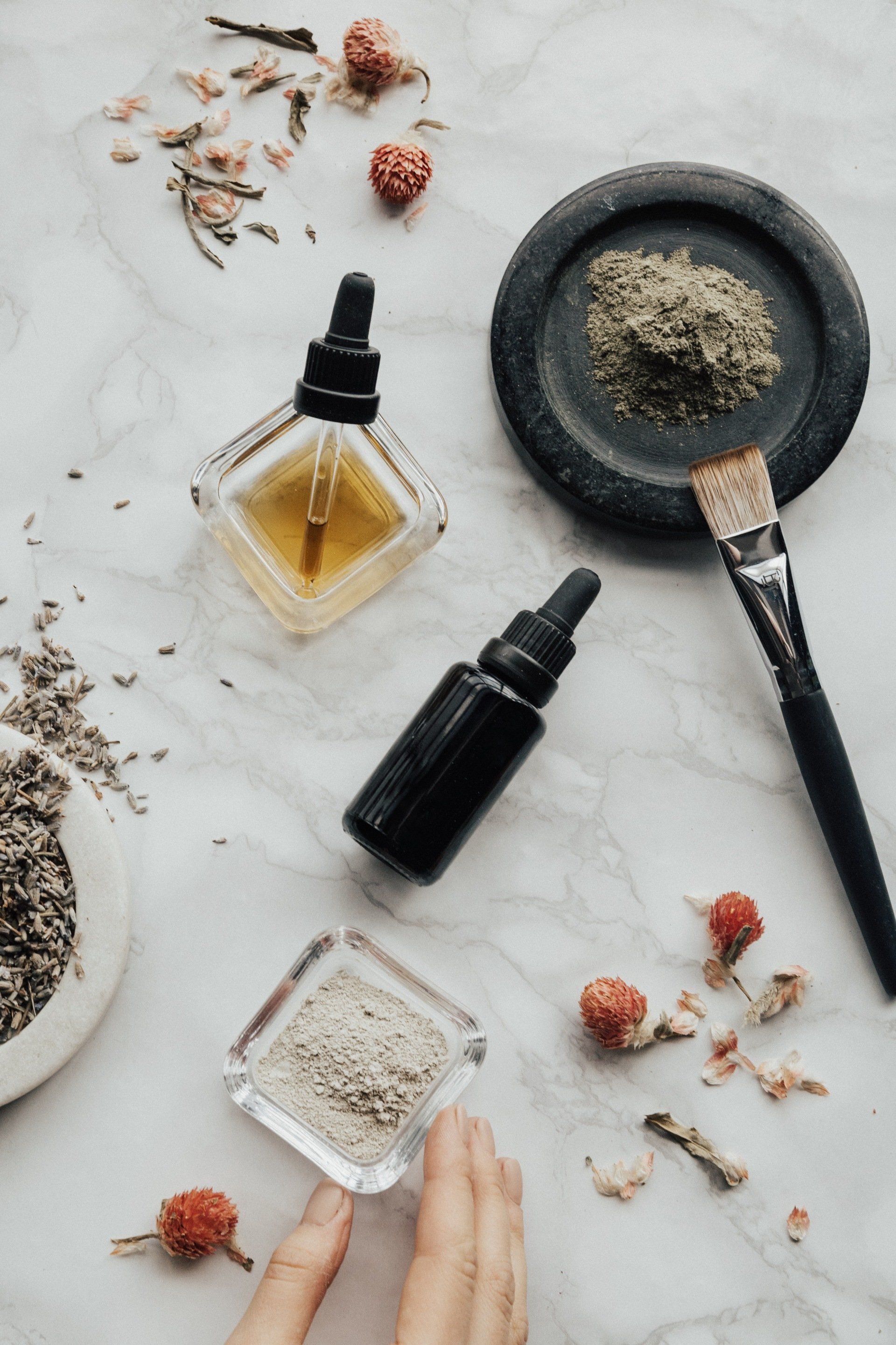 A high-angle shot shows two essential oil bottles, facial mask powders, a brush, and dried botanicals on a marble surface.