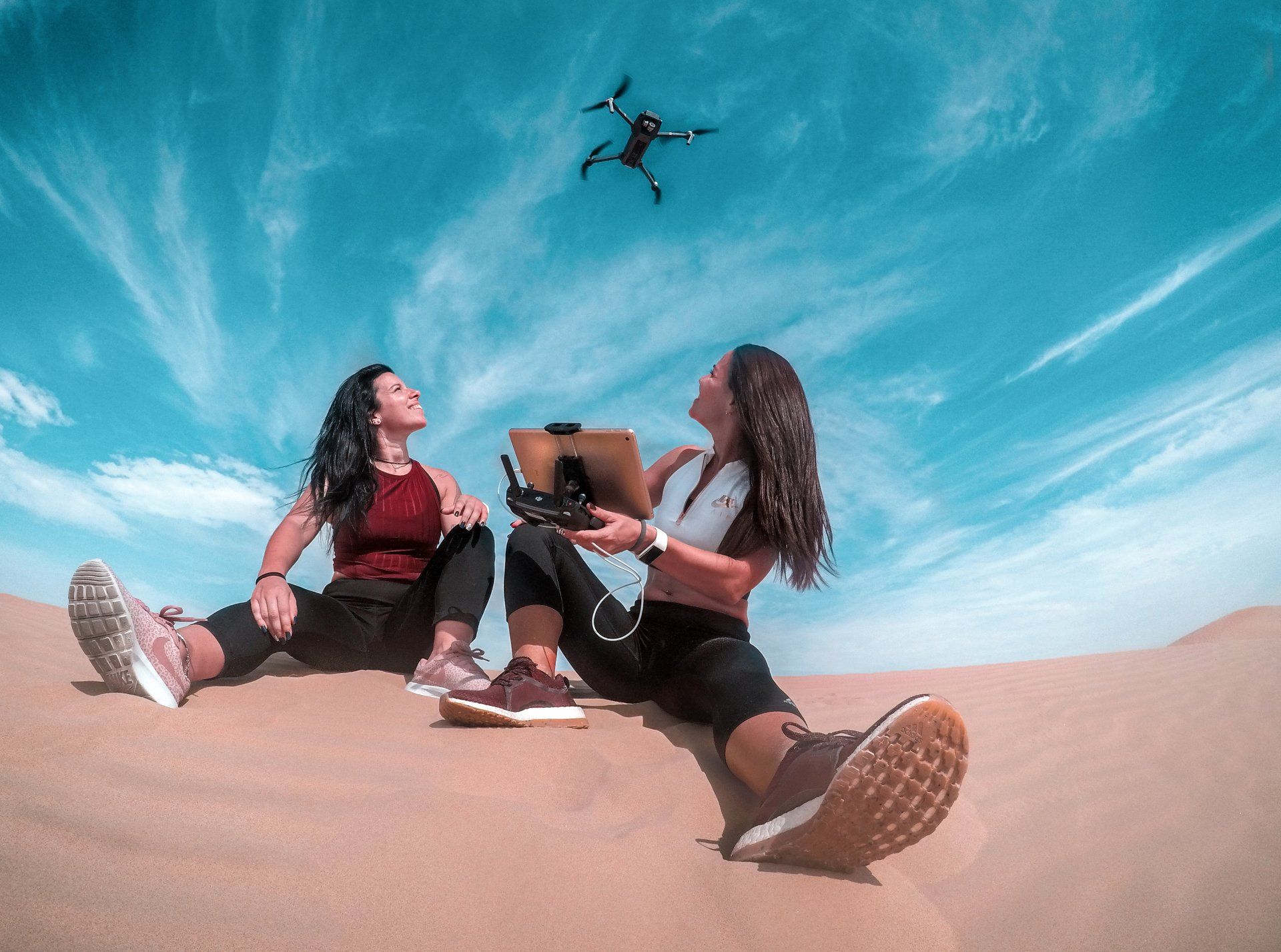 Duas mulheres estão sentadas no topo de uma duna de areia com um drone voando ao fundo.