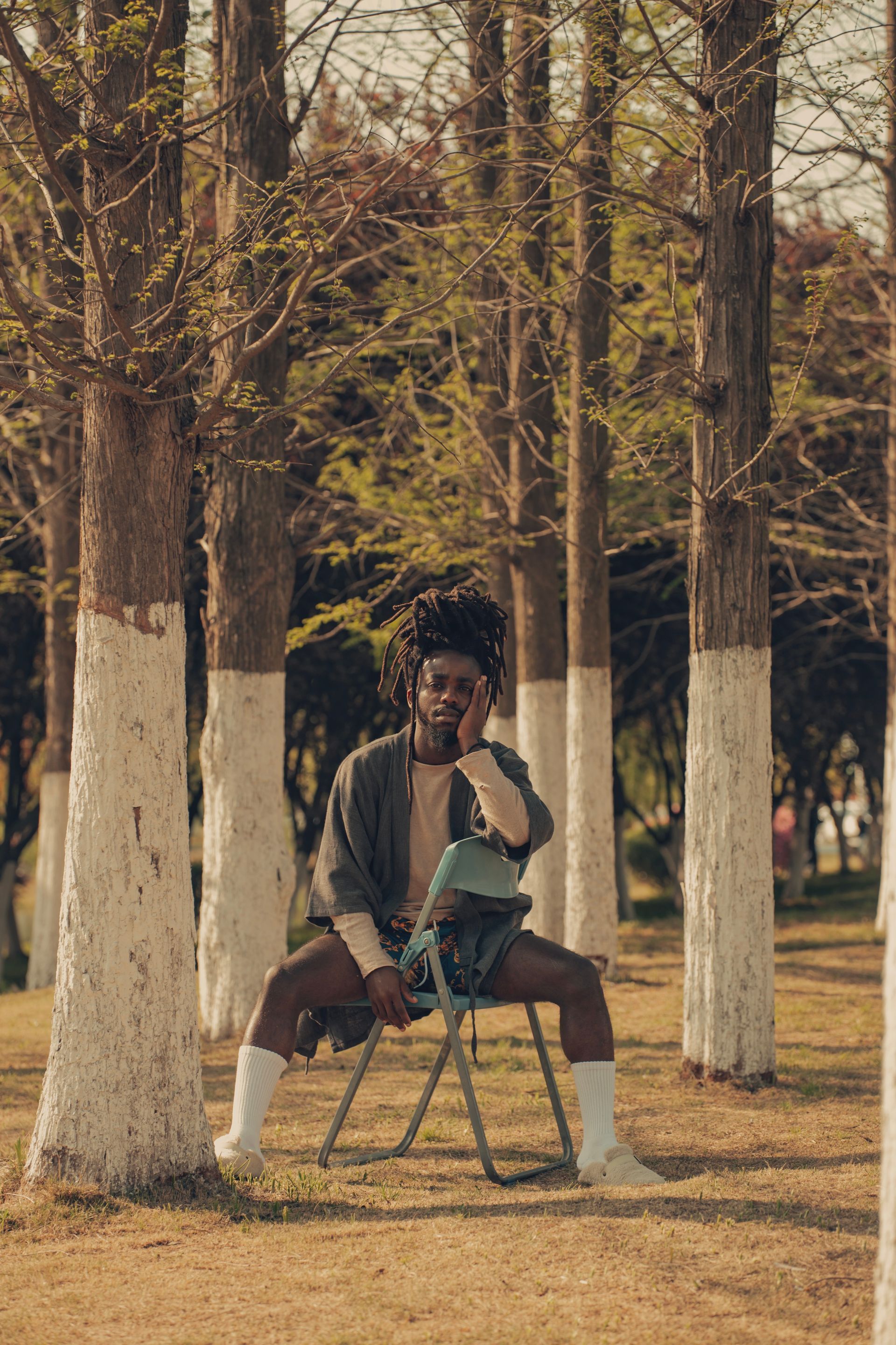 Person sits on a chair in a tree-filled park, wearing a jacket and white socks, looking thoughtful.