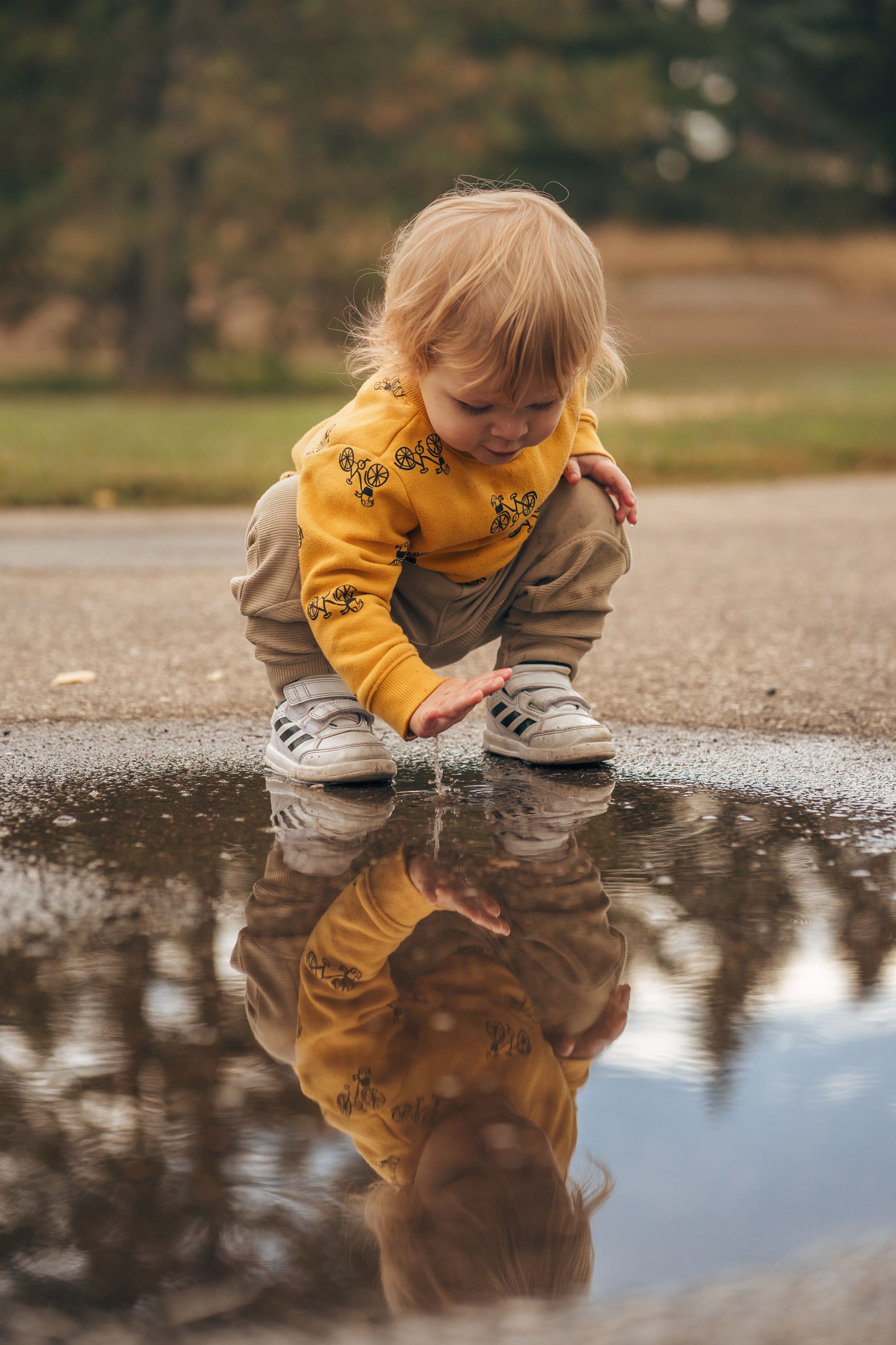 Child squatting, touching a puddle, reflected in the water. Blonde hair, yellow sweater, gray pants, outdoors.