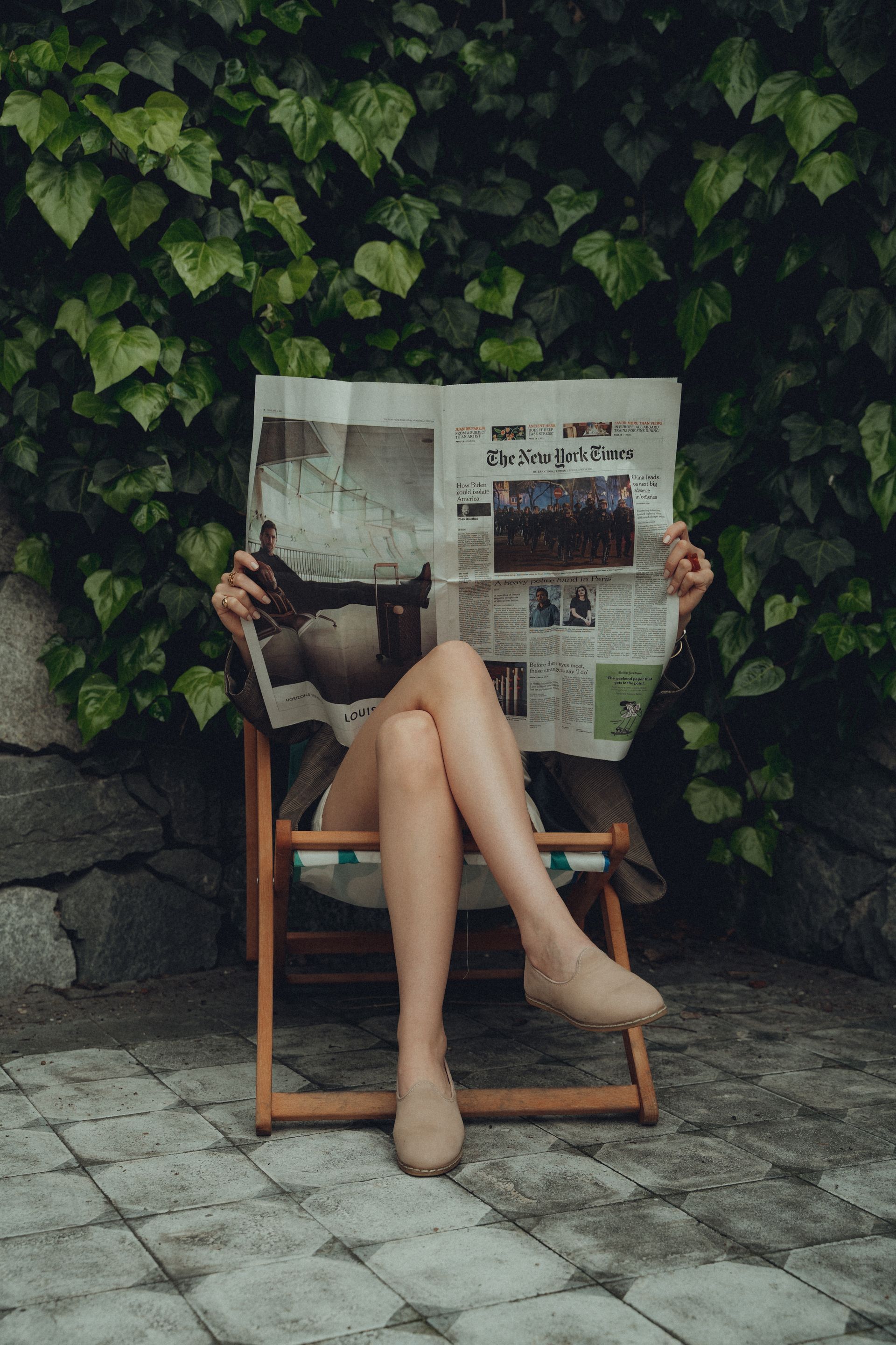 Een persoon leest buiten de krant, zittend op een houten stoel met de benen gekruist. Groene klimplanten op de achtergrond.