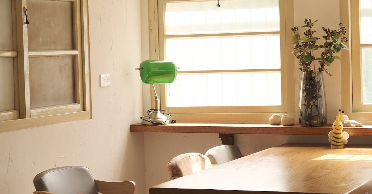 Bright dining room with a wooden table, green desk lamp, and vase of flowers by a sunlit window.