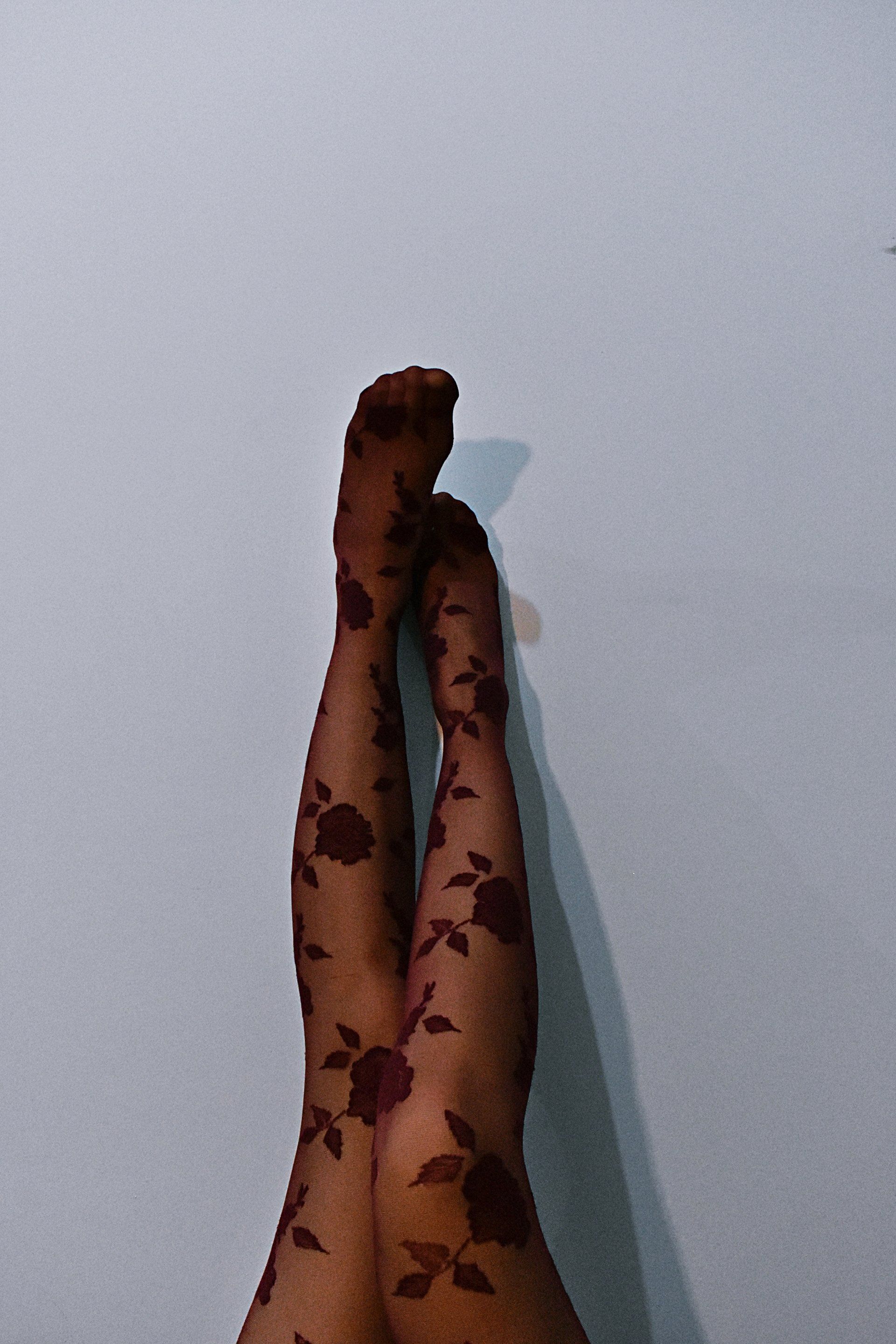 Gambe in collant trasparenti marroni con motivo floreale, appoggiate contro un muro bianco.