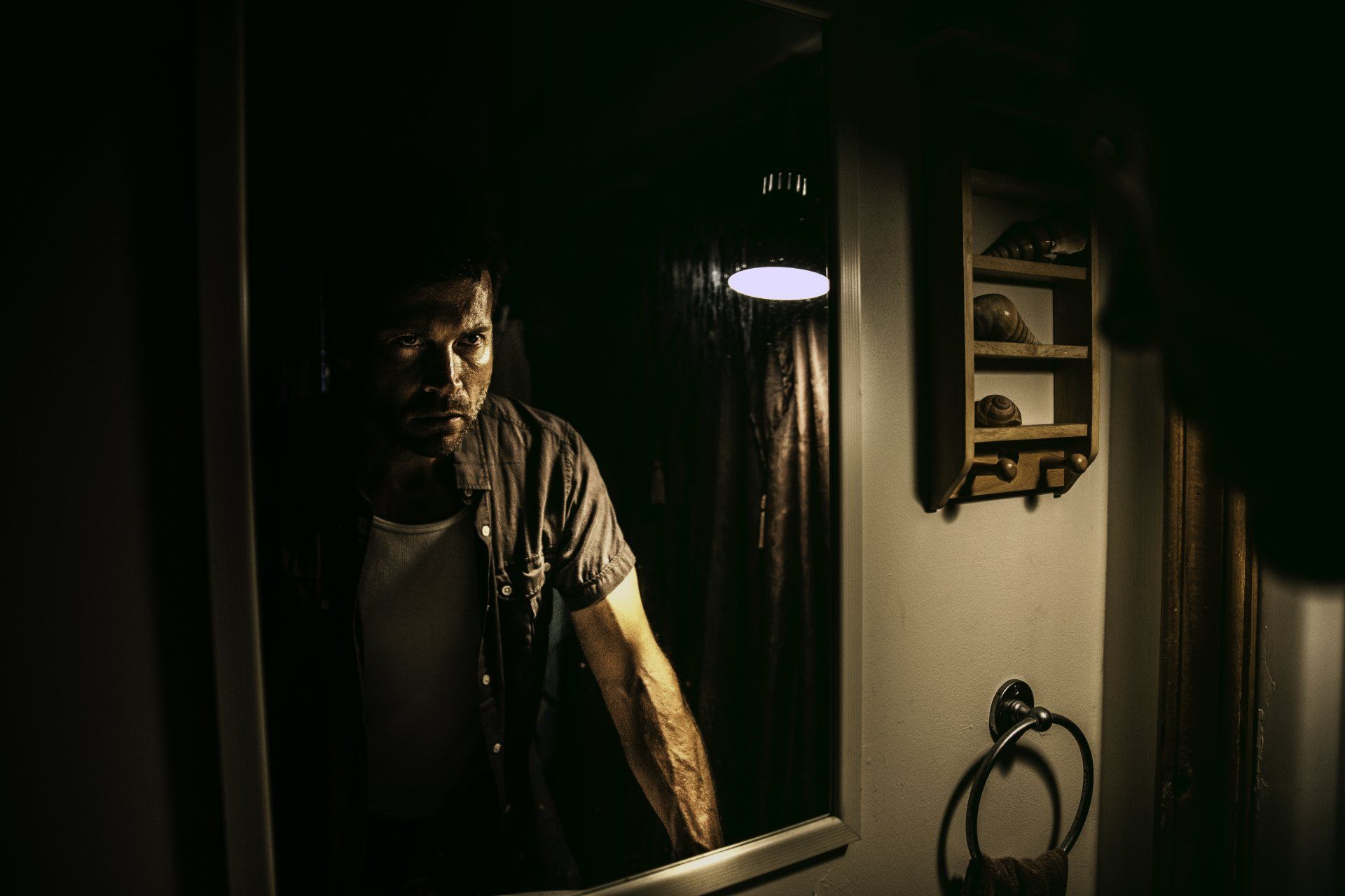 Man leaning in a dim hallway mirror, lit by a single overhead lamp, with a towel and shelf nearby