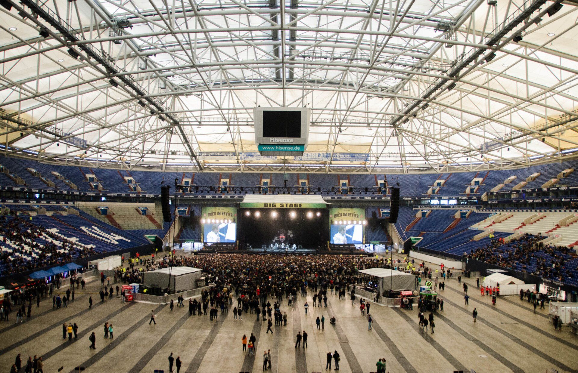 Large concert inside a stadium. Stage setup with band performing. Crowd gathered in front.