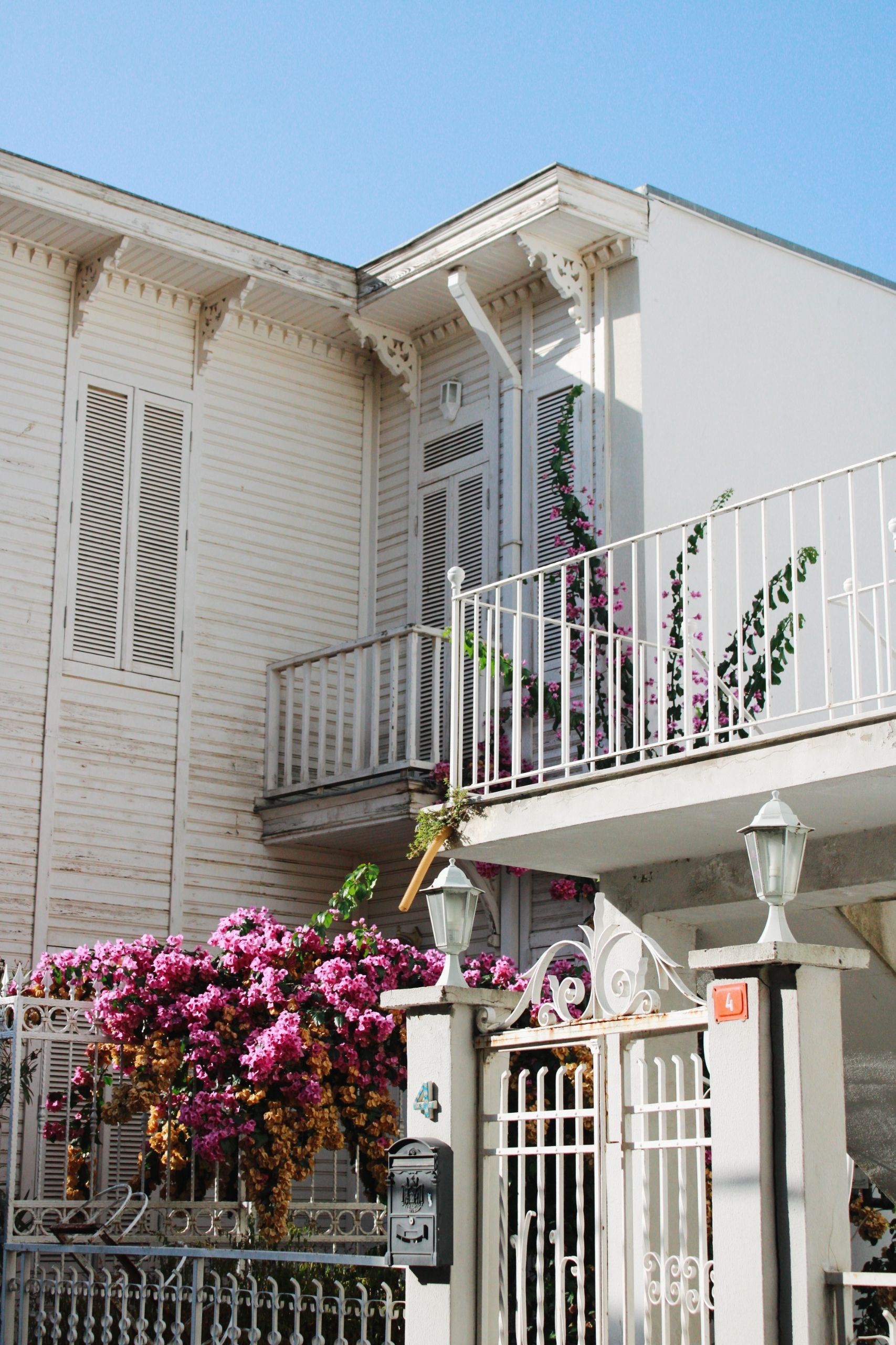 White house with balcony, pink flowers, and an ornate front gate under a clear blue sky