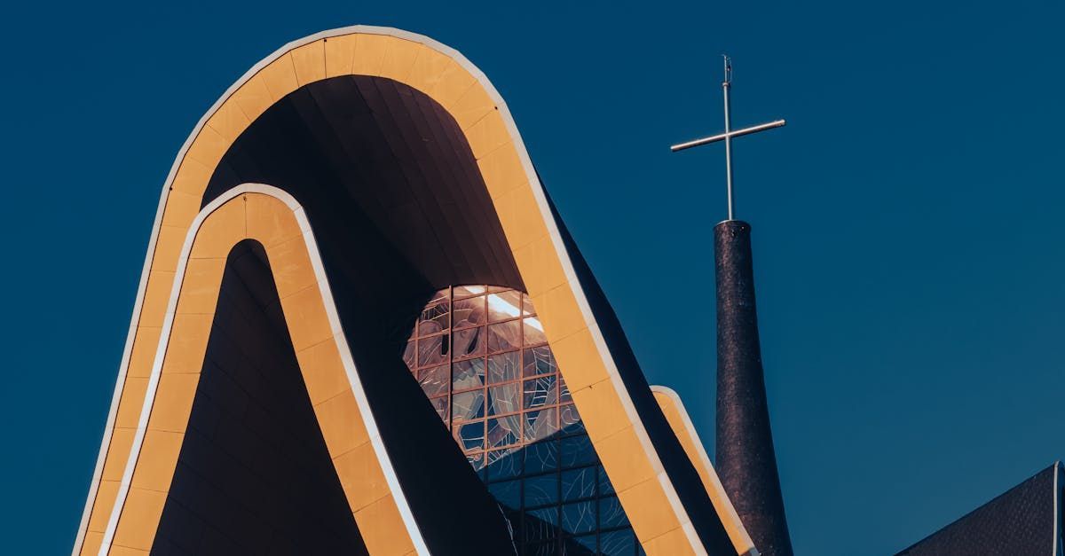 Curved orange architectural sculpture against a blue sky with a cross atop a dark tower in the background