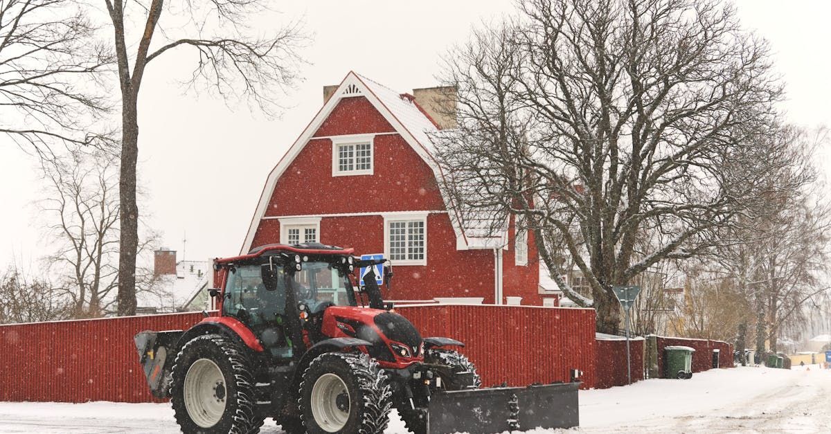Red tractor in snowy yard beside a red house and bare trees, with snow falling.