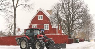 Red tractor in snowy yard beside a red house and bare trees, with snow falling.