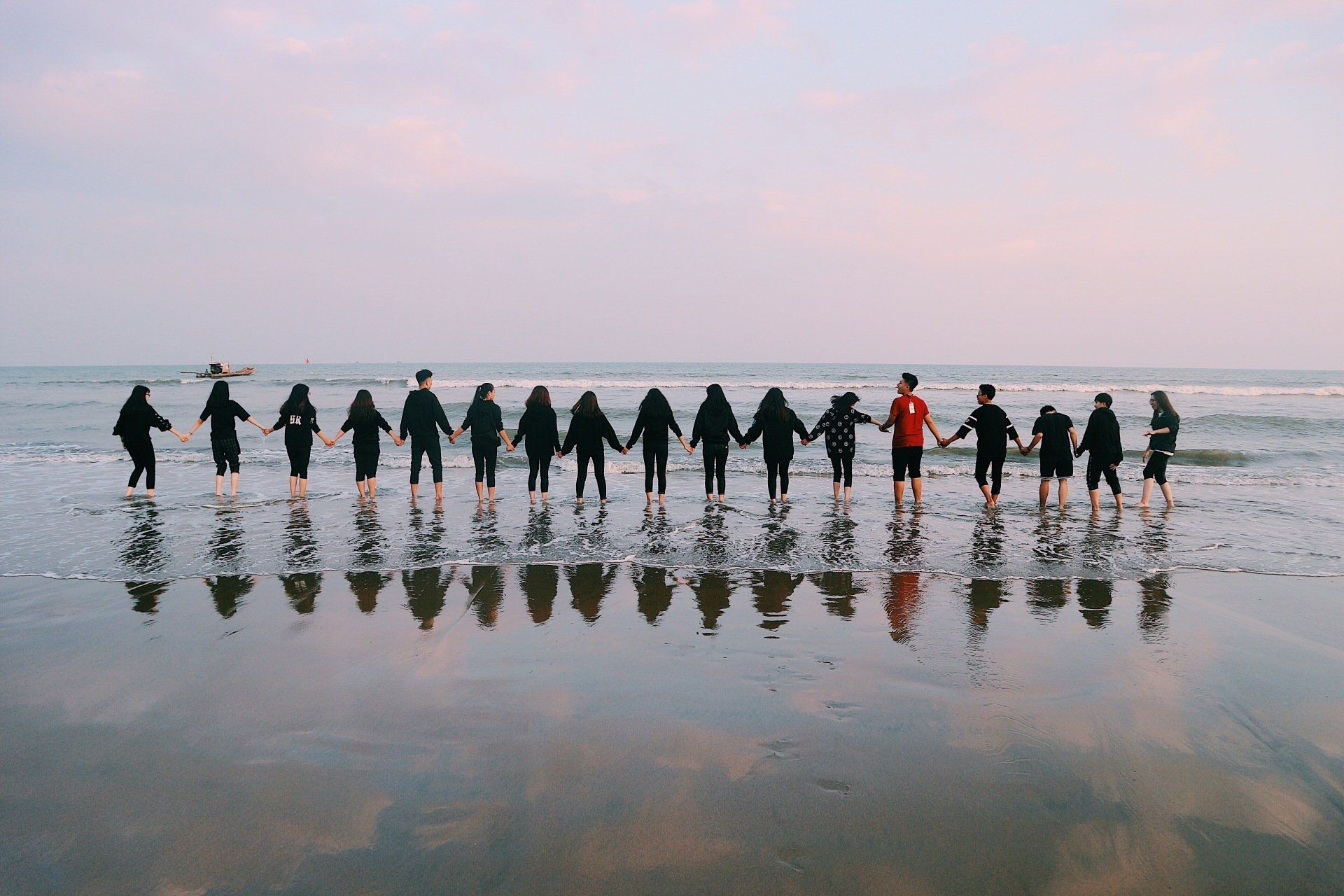 a long line of people all in black, with one person in red with their backs to the camera looking out to the sea. they are on the edge of the waves with their feet in the water