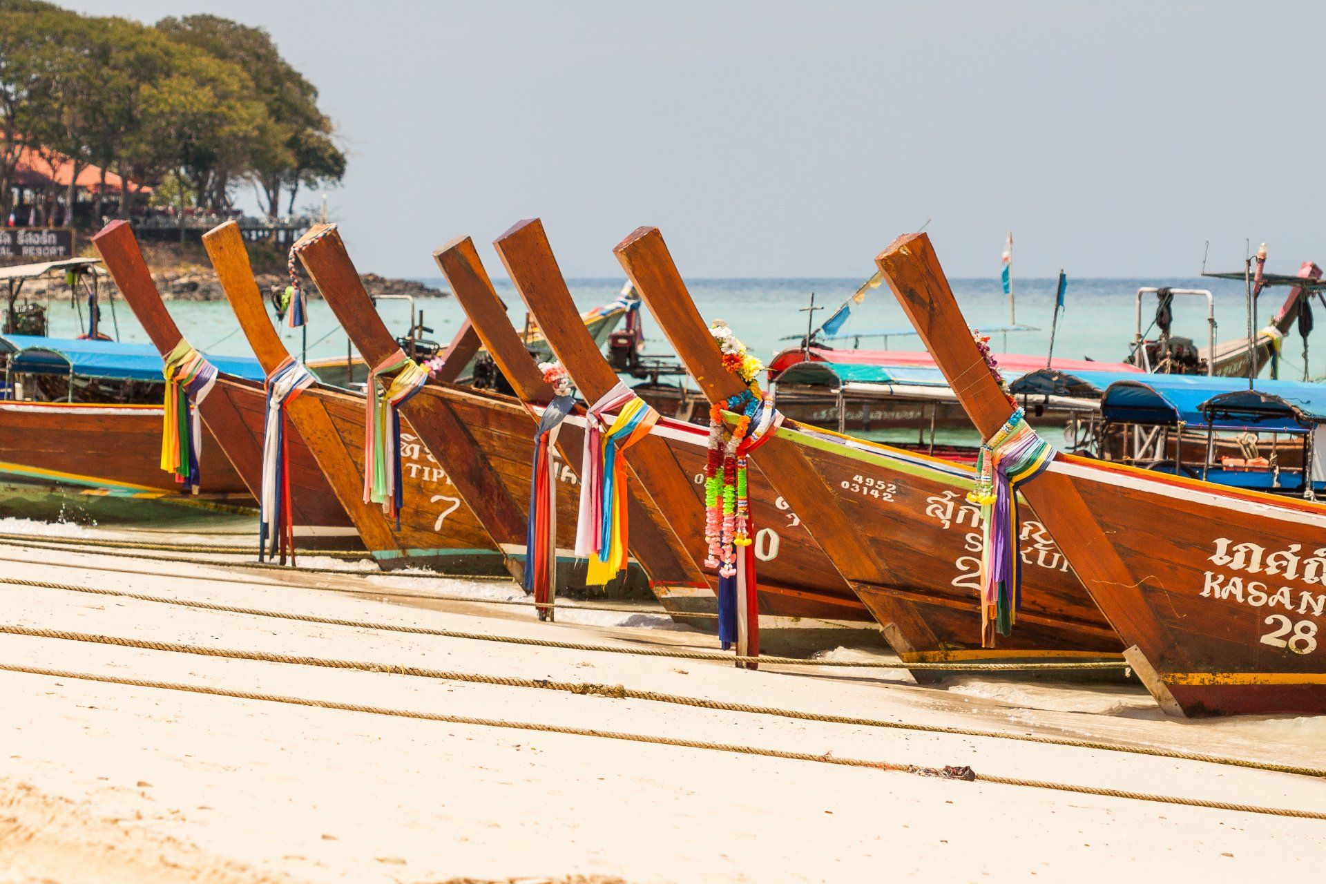 Longtail boats lined up on a beach in Thailand, with colorful ribbons on the prows.