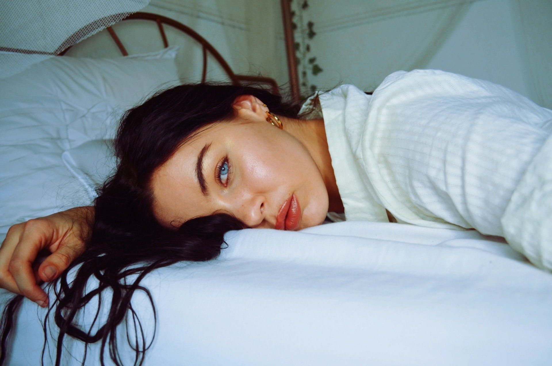 Woman lying on a white bed, wearing a robe, looking towards the camera with a relaxed expression.