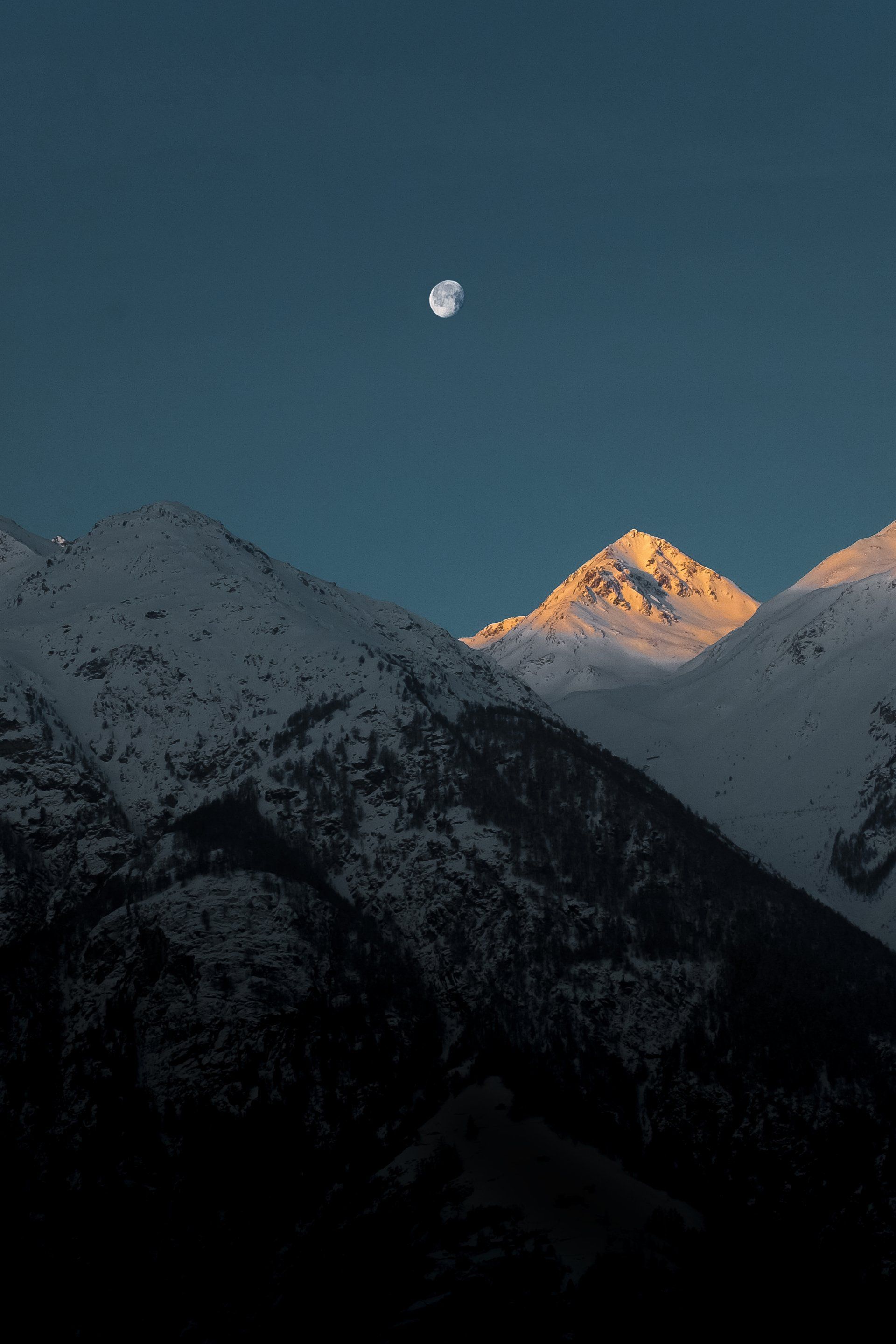 Snowy mountains at dusk with moon in the blue sky. The peak is illuminated by sunlight.