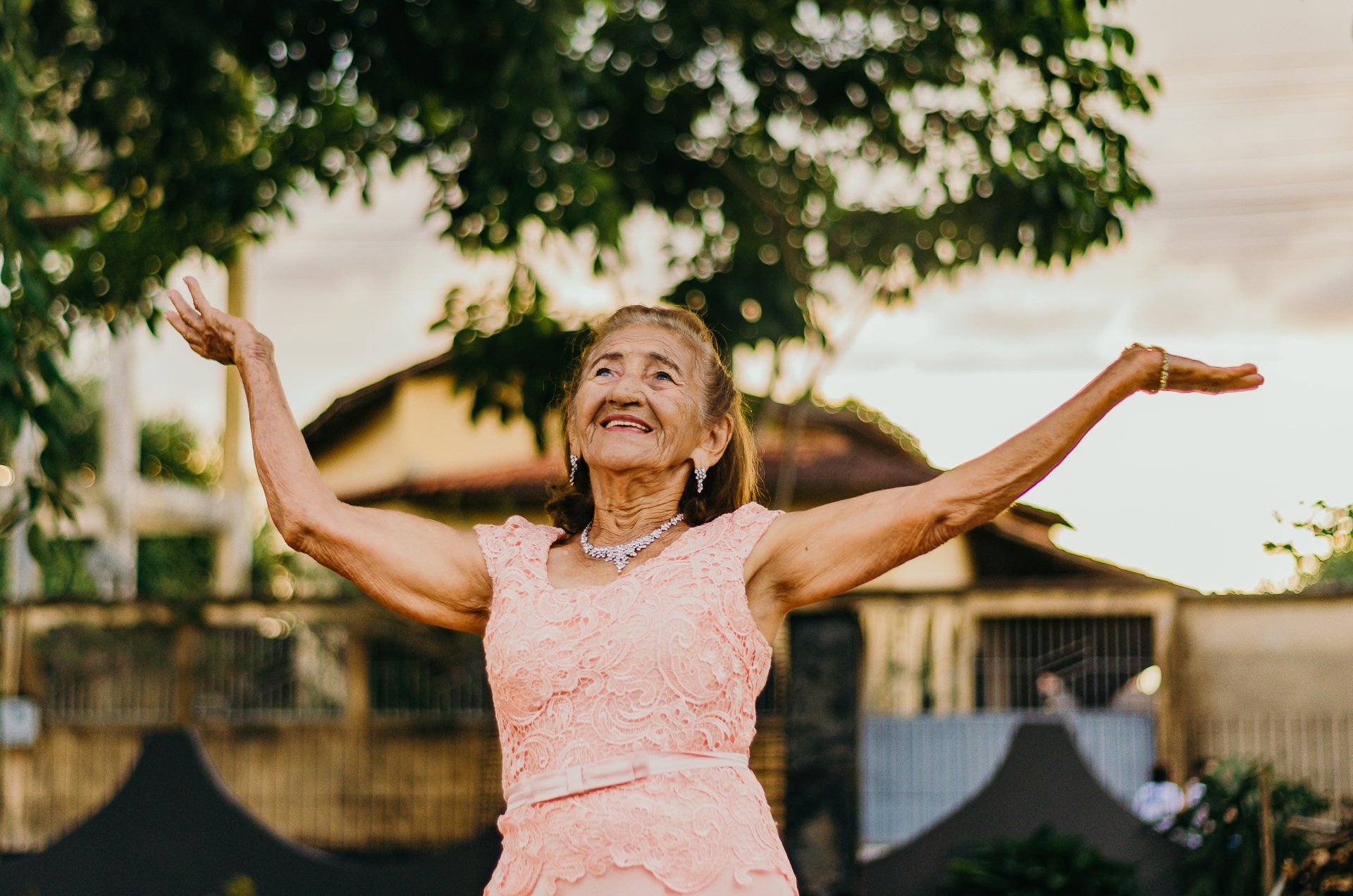 An elderly woman in a pink dress is dancing with her arms outstretched.