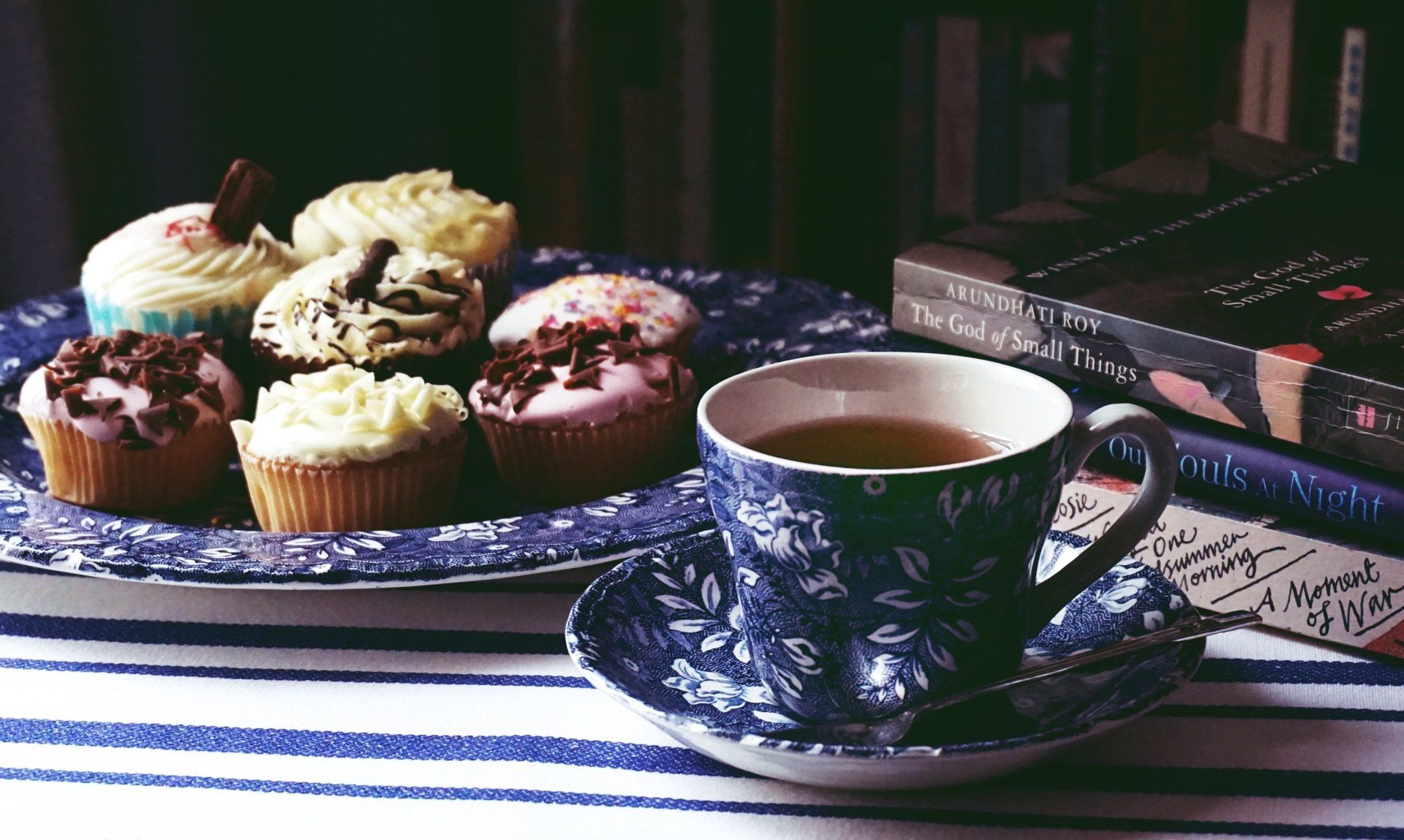 Cup of tea and cupcakes on a floral plate, beside books on a striped tablecloth.