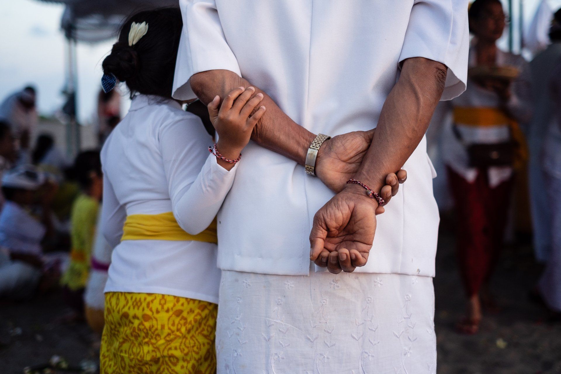 A child in white and yellow traditional attire holds the arm of an adult wearing white, both standing in a crowd.