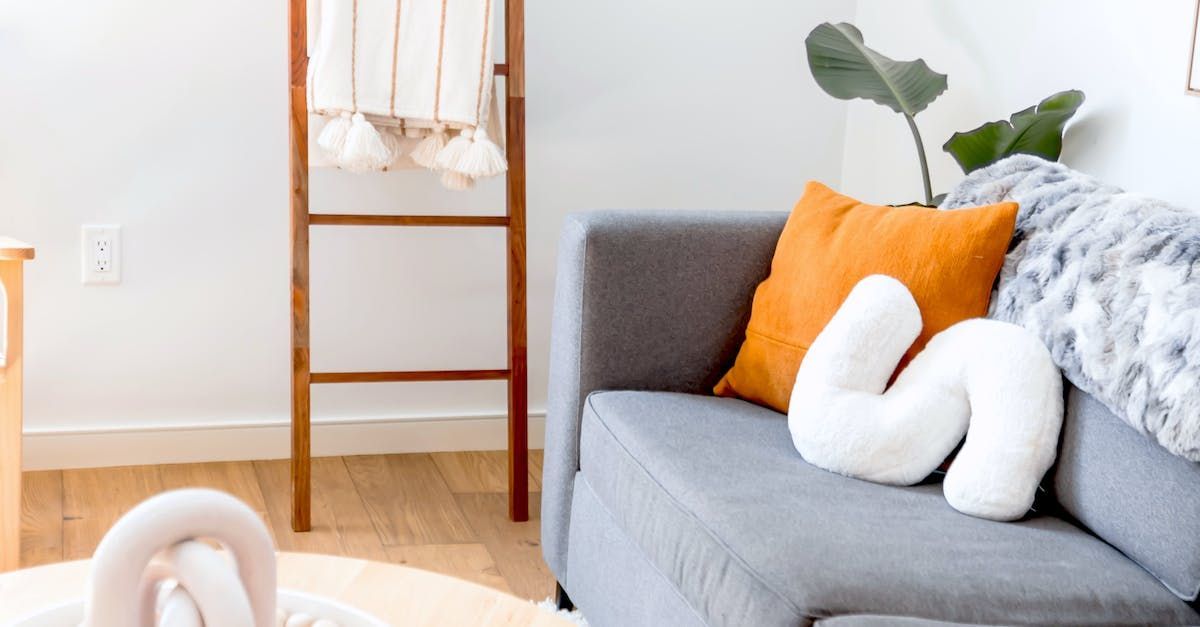 A grey couch with orange and white decorative pillows, next to a wooden ladder with a blanket in a brightly lit room.